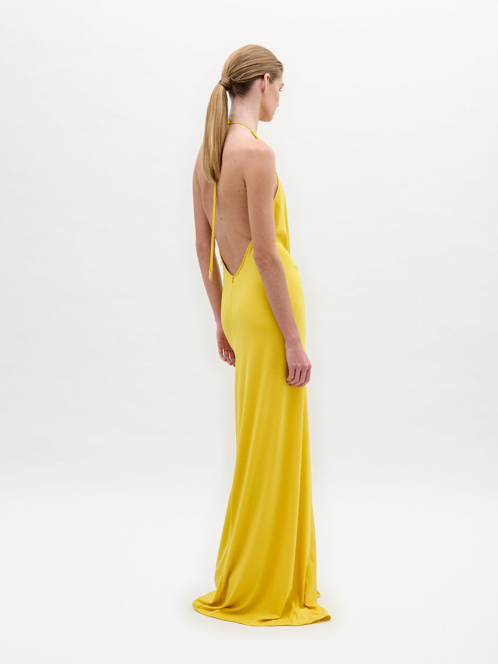 Someone stands in an Aiko Dress, a long yellow halter style with a circular design at the waist, set against a plain white background.