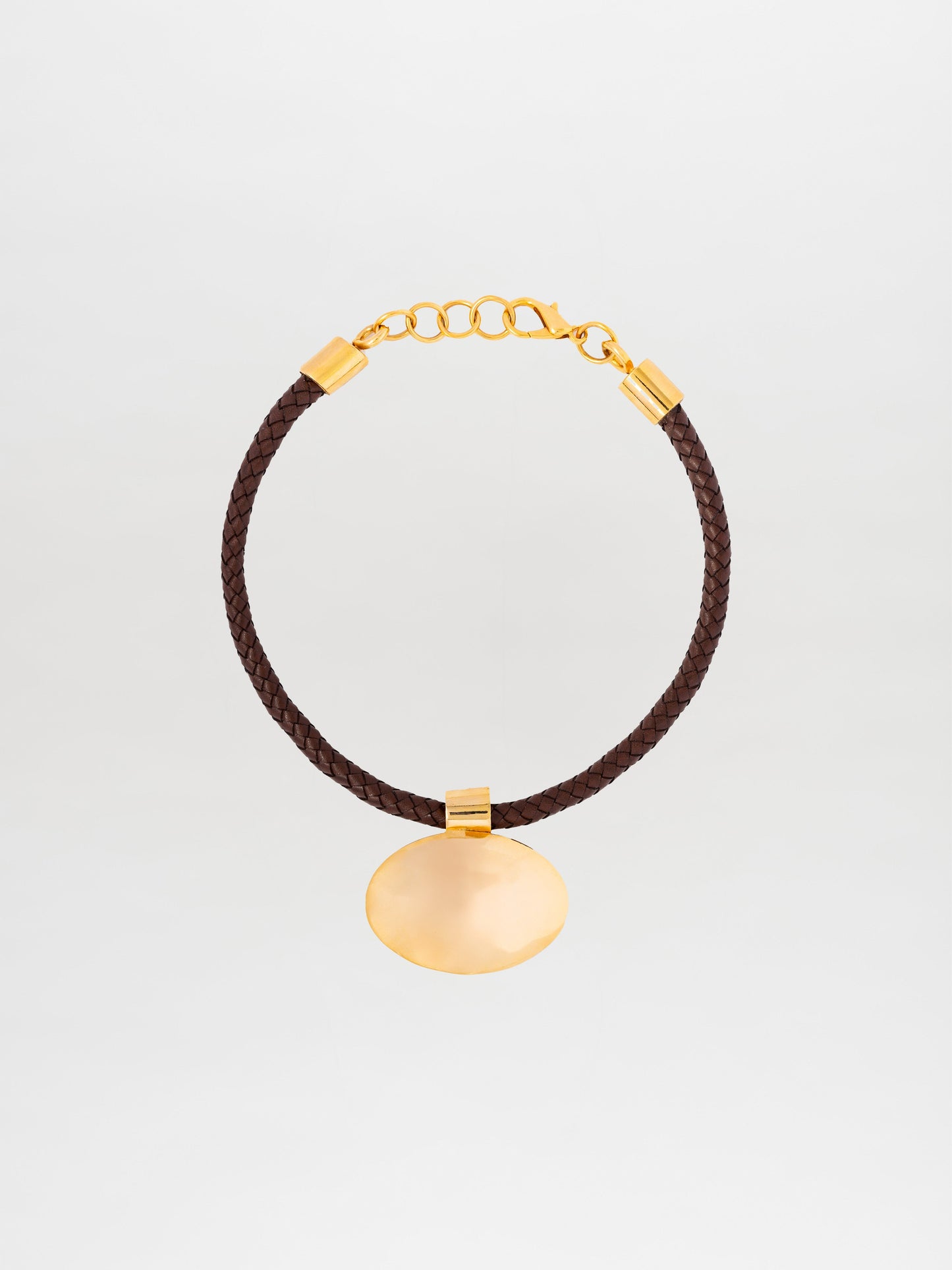 El Collar Gimena es una gargantilla de cuero trenzado marrón con cadena y broche de oro, elaborada en Colombia. Presenta un gran colgante ovalado de oro en el centro, sobre un fondo liso de color claro.
