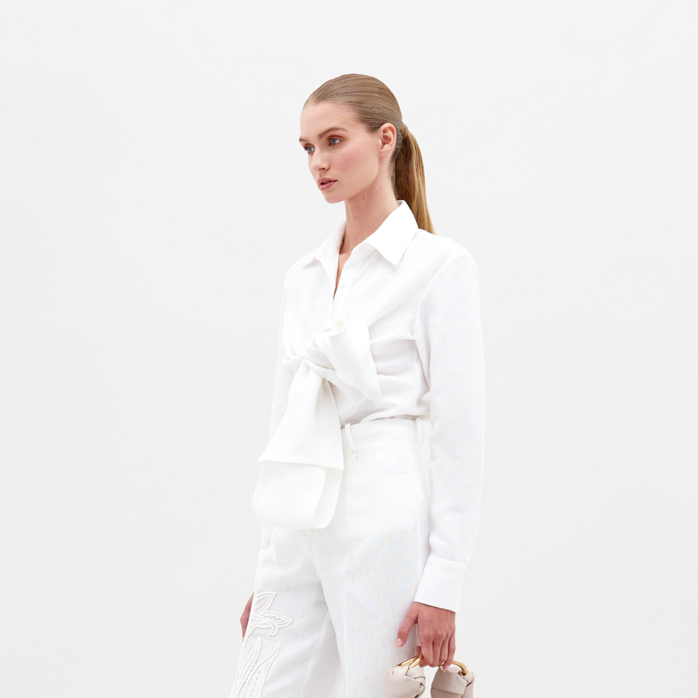 A person with long light brown hair in a ponytail is wearing the Mercury Blouse (Linen) White with a large bow, white pants, and holding beige shoes. Pre-order now to ensure you get this look by the 2025 ship-by date.