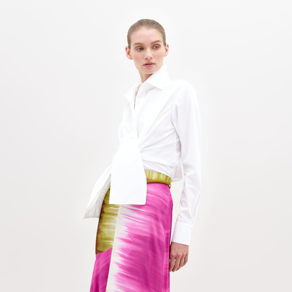 Una persona con el cabello recogido viste la Blusa Mercury, una blusa blanca de manga larga abotonada con un dobladillo asimétrico, combinada con una falda colorida con vibrantes textiles portugueses en rosa, blanco y verde sobre un fondo blanco liso.