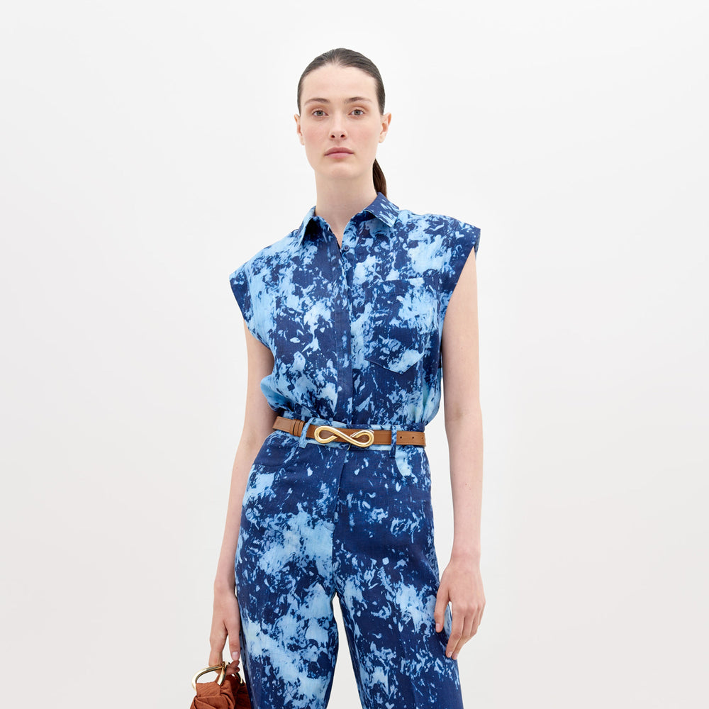Una persona está parada sobre un fondo blanco vistiendo la Blusa Rina, un mono azul sin mangas con efecto tie-dye y estampado abstracto, y sostiene un bolso marrón en la mano derecha. Lleva un cinturón marrón con una hebilla ovalada y el cabello recogido hacia atrás con cuidado.