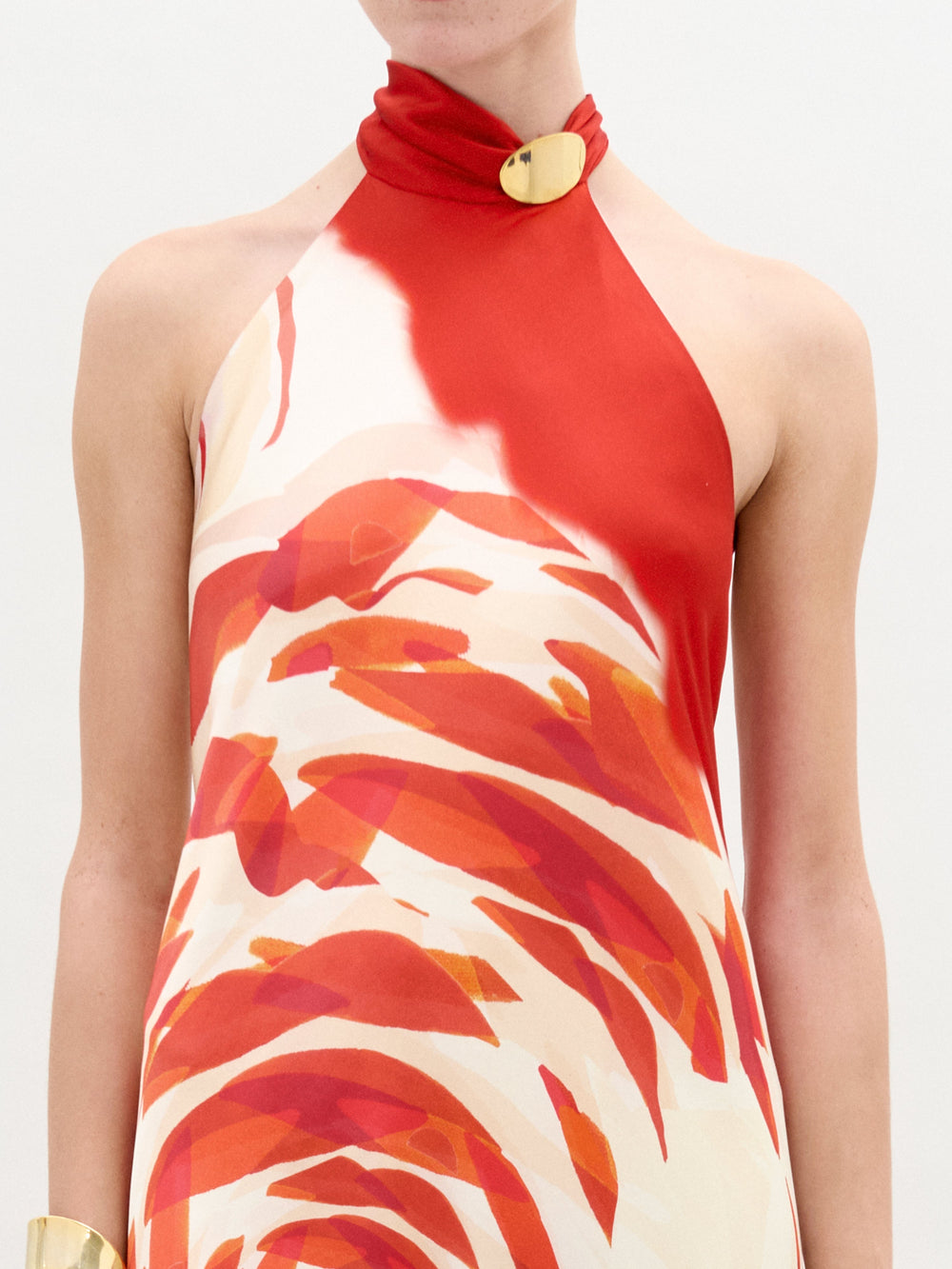 Alguien lleva el Vestido Sherry, un vestido de seda sin mangas con escote halter que presenta un patrón de remolinos en rojo y naranja sobre fondo blanco. Tiene un broche dorado en el cuello, combinado con parte de una pulsera dorada visible en la muñeca izquierda.