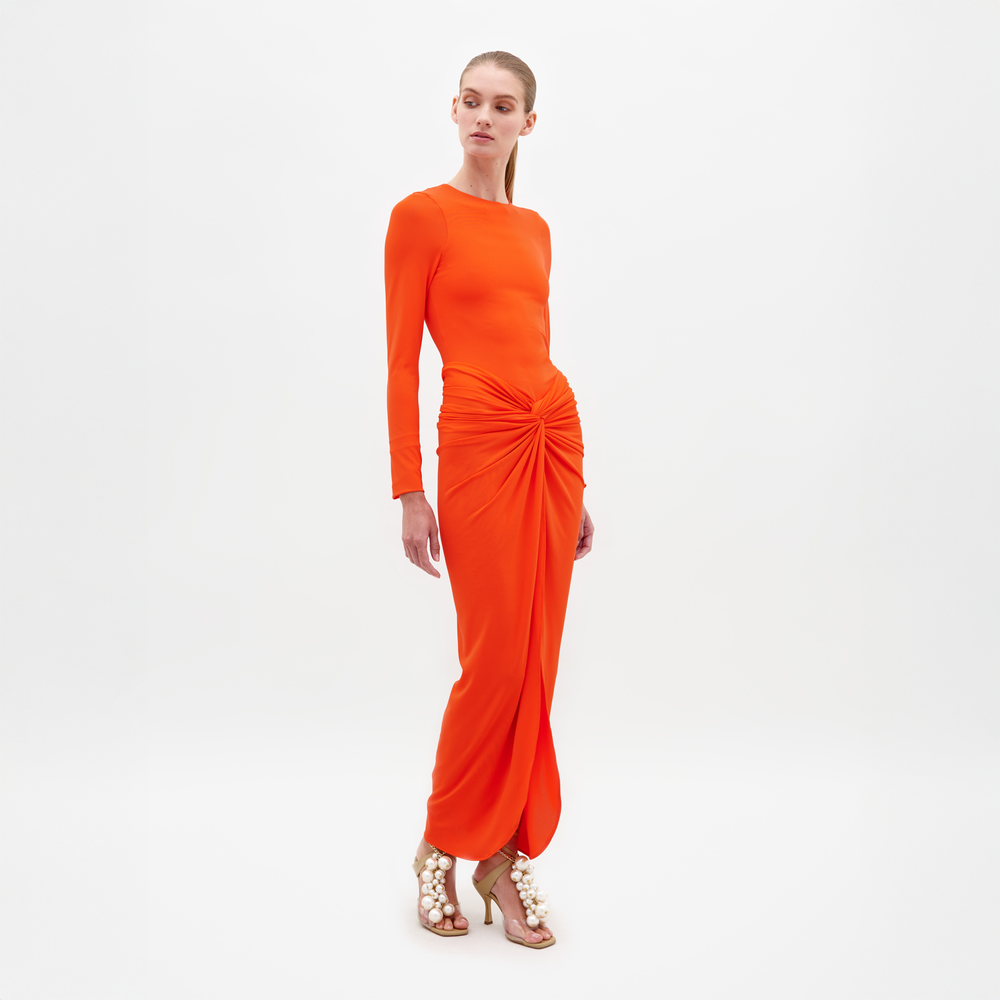 En un estudio, una persona luce el vestido Sydelle, un maxivestido naranja vibrante de manga larga con un nudo en la cintura. Sus tacones altos abiertos presentan elementos decorativos. Un fondo blanco resalta la elegancia del conjunto.