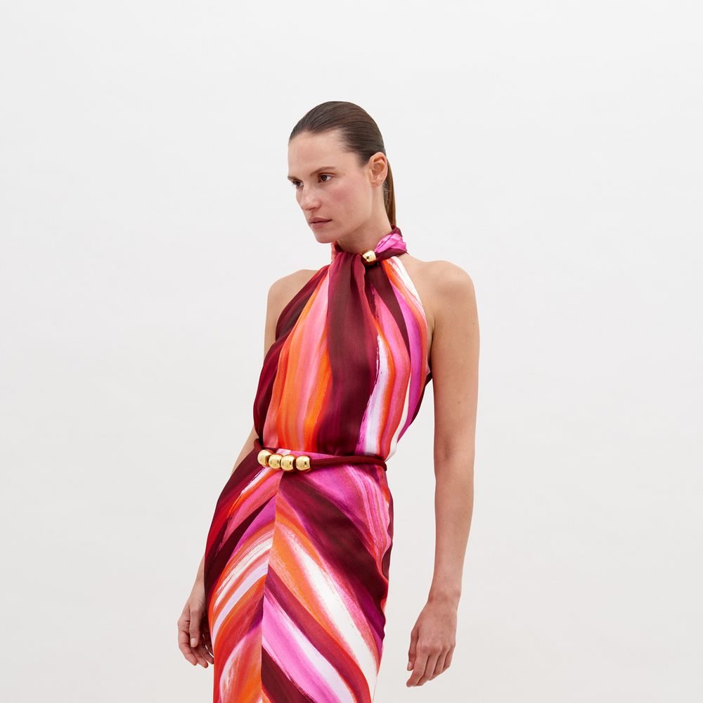 A woman stands against a plain background, wearing a halter-neck dress with a multicolored pattern of pink, orange, and red hues, accessorized with a gold belt buckle. Pre-order the Shela Blouse Magenta Abstract Waves now to ensure delivery by November 15th, 2024.