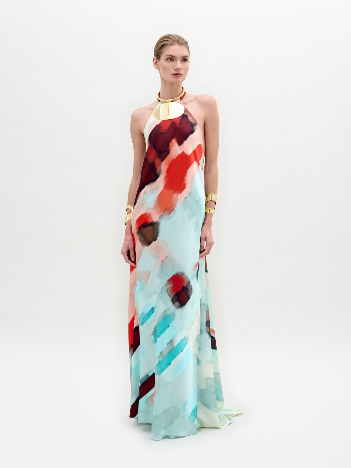 Una persona lleva el Vestido Zeffa, un vestido largo de seda con un estampado abstracto en rojo, azul y blanco. Tiene un escote halter y está decorado con brazaletes y un collar dorados sobre un fondo blanco liso.