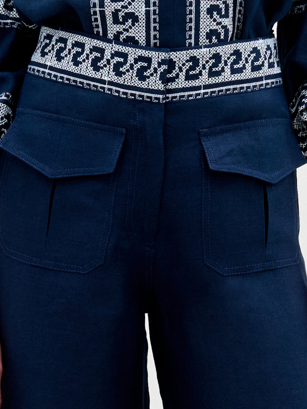 Primer plano de una persona que lleva el Pantalón Zina en azul oscuro, de cintura alta y dos grandes bolsillos delanteros. El estilo de pierna ancha se complementa con un cinturón estampado y un top a juego con intrincados diseños en azul y blanco, todo sobre un fondo en tonos neutros que realza la elegancia del conjunto.