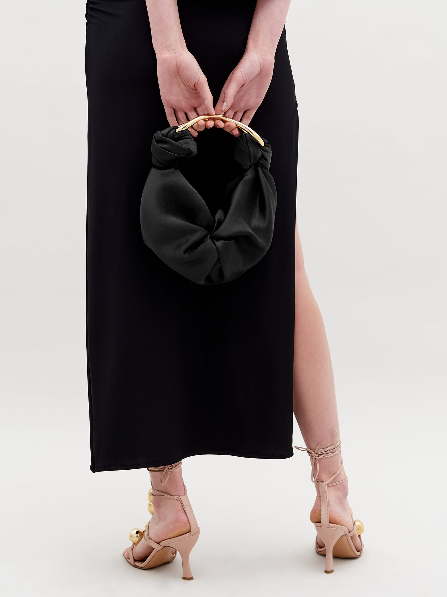 Una persona con un vestido negro sostiene el Bolso Sofia Small, un pequeño bolso negro con asa dorada y un dije con la leyenda "Hecho en Colombia", detrás de su espalda. Lleva unas sandalias de tacón alto color beige sobre un fondo blanco liso.