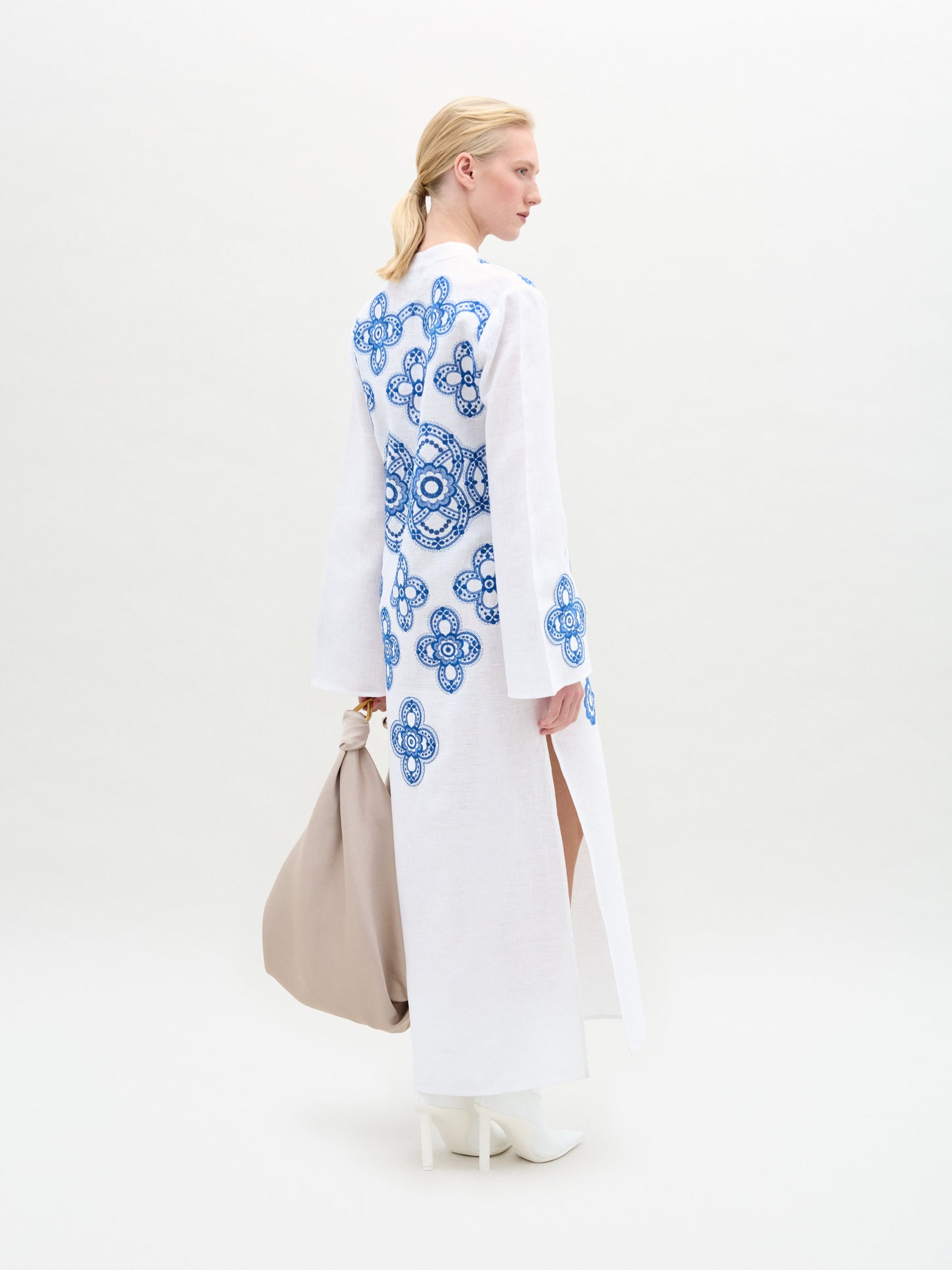 Dressed in the Adalys Tunic, featuring blue patterns on white fabric, and holding a beige bag while wearing white boots, a person stands against a plain background.