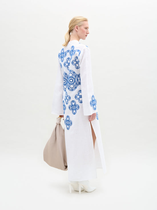 Dressed in the Adalys Tunic, featuring blue patterns on white fabric, and holding a beige bag while wearing white boots, a person stands against a plain background.