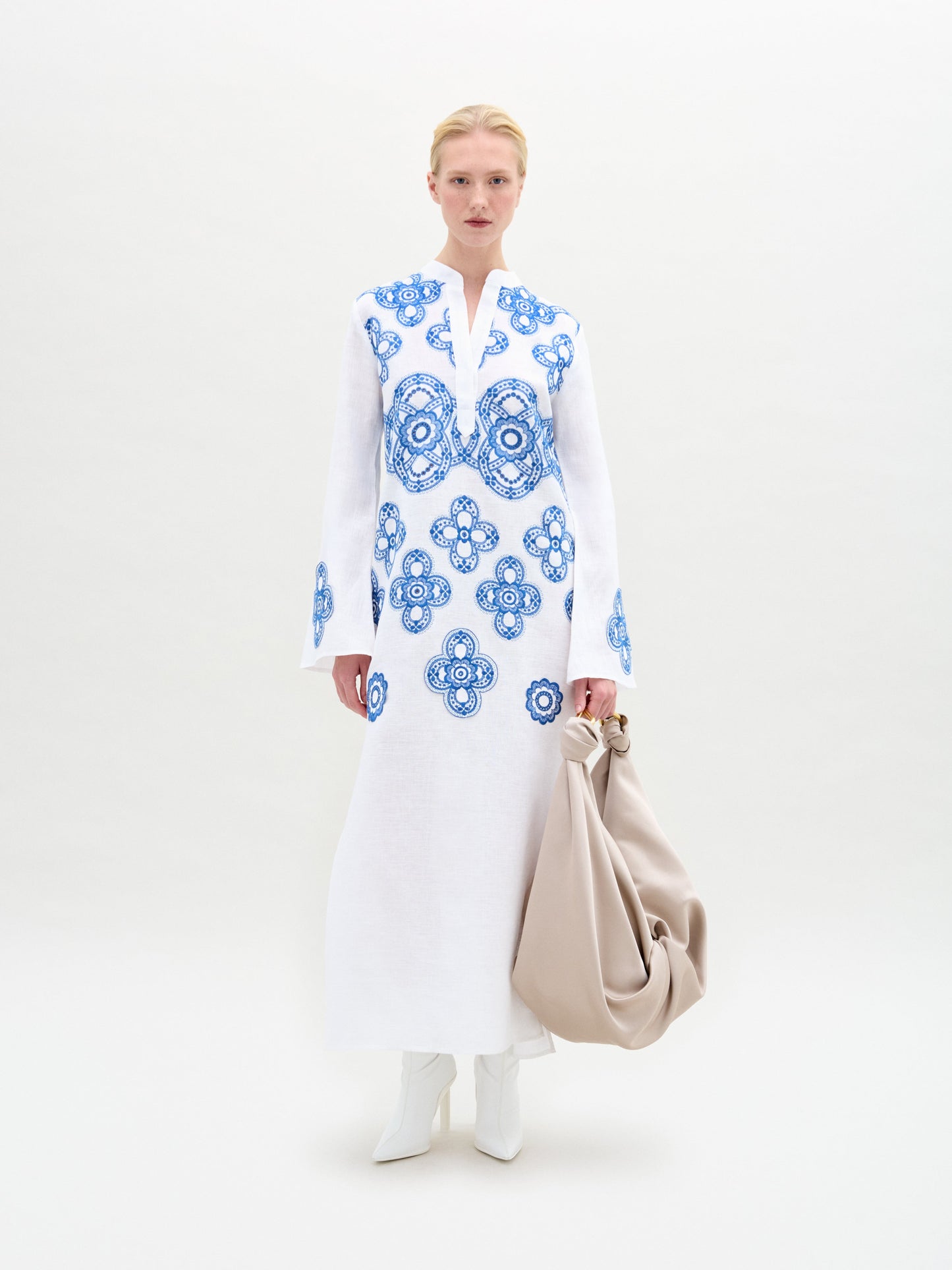 Dressed in the Adalys Tunic, featuring blue patterns on white fabric, and holding a beige bag while wearing white boots, a person stands against a plain background.