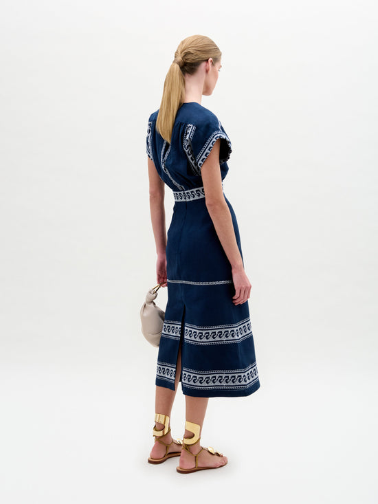 Una mujer rubia con cola de caballo está parada de espaldas a la cámara, luciendo el Vestido Ady azul adornado con patrones blancos. Sus sandalias doradas y su pequeño bolso beige completan su look, resaltando su exquisito diseño hecho en Colombia sobre un fondo blanco liso.