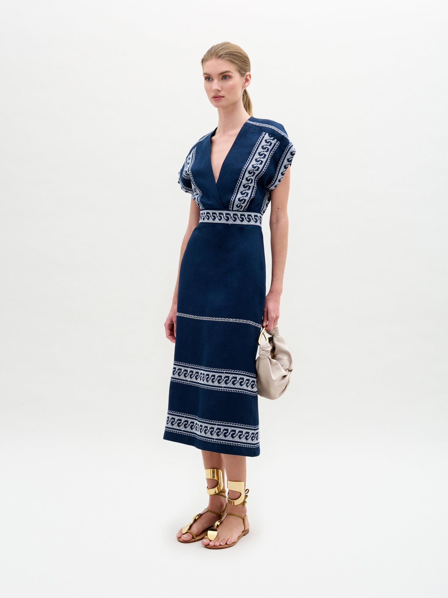 A woman with blonde hair in a ponytail is seen from the back wearing the Ady Dress, featuring a blue pattern, styled with sandals and holding a beige bag against a plain background.
