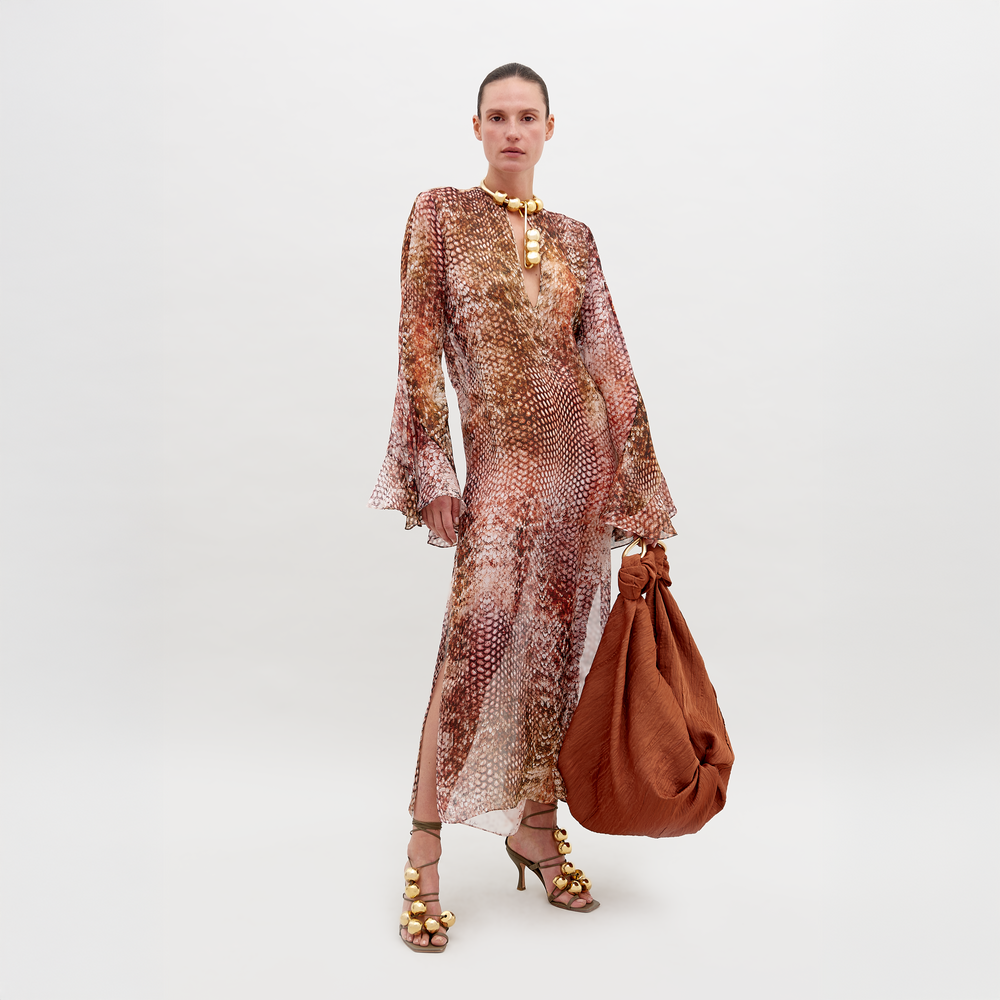 Frente a un fondo liso, una persona lleva el Vestido Aries, un vestido de seda largo y vaporoso con escote alto y mangas acampanadas. Sostiene un gran bolso de color óxido y calza sandalias de tiras doradas, mirando directamente a la cámara.