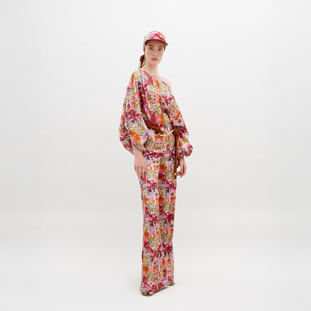 Una persona está parada sobre un fondo liso con Pantalón Andie, un conjunto con estampado floral que incluye una blusa con mangas voluminosas, pantalones bota ancha de cintura alta a juego y una gorra. El conjunto combina rosa, amarillo y otros colores.