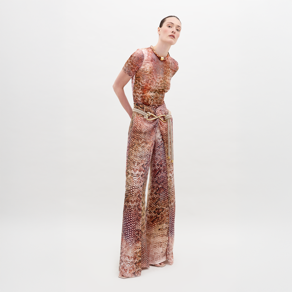 Una persona lleva el Pantalón Canturipe, de pierna ancha y cintura alta, combinado con un top estampado en tela italiana marrón y rosa. Agrega un cinturón y un collar dorados mientras se para elegantemente sobre un fondo blanco liso.