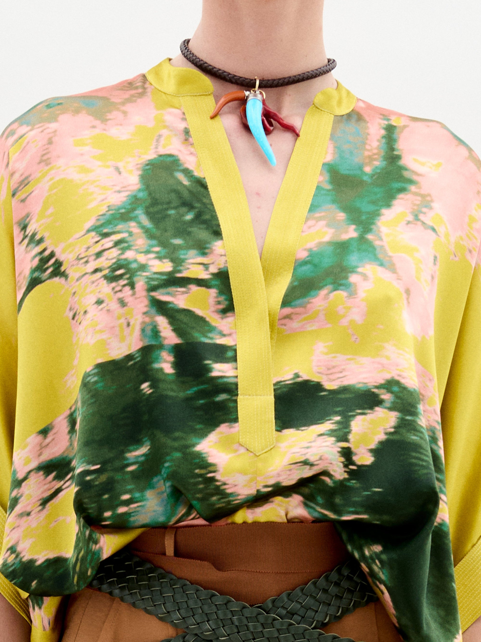 Una persona lleva la Blusa Daia, una lujosa blusa de tela italiana con estampados abstractos en verde, amarillo y rosa vibrantes. La blusa suelta con cuello en V se combina con un cinturón trenzado marrón, mientras que un collar con colgantes azules y naranjas agrega el toque final perfecto.