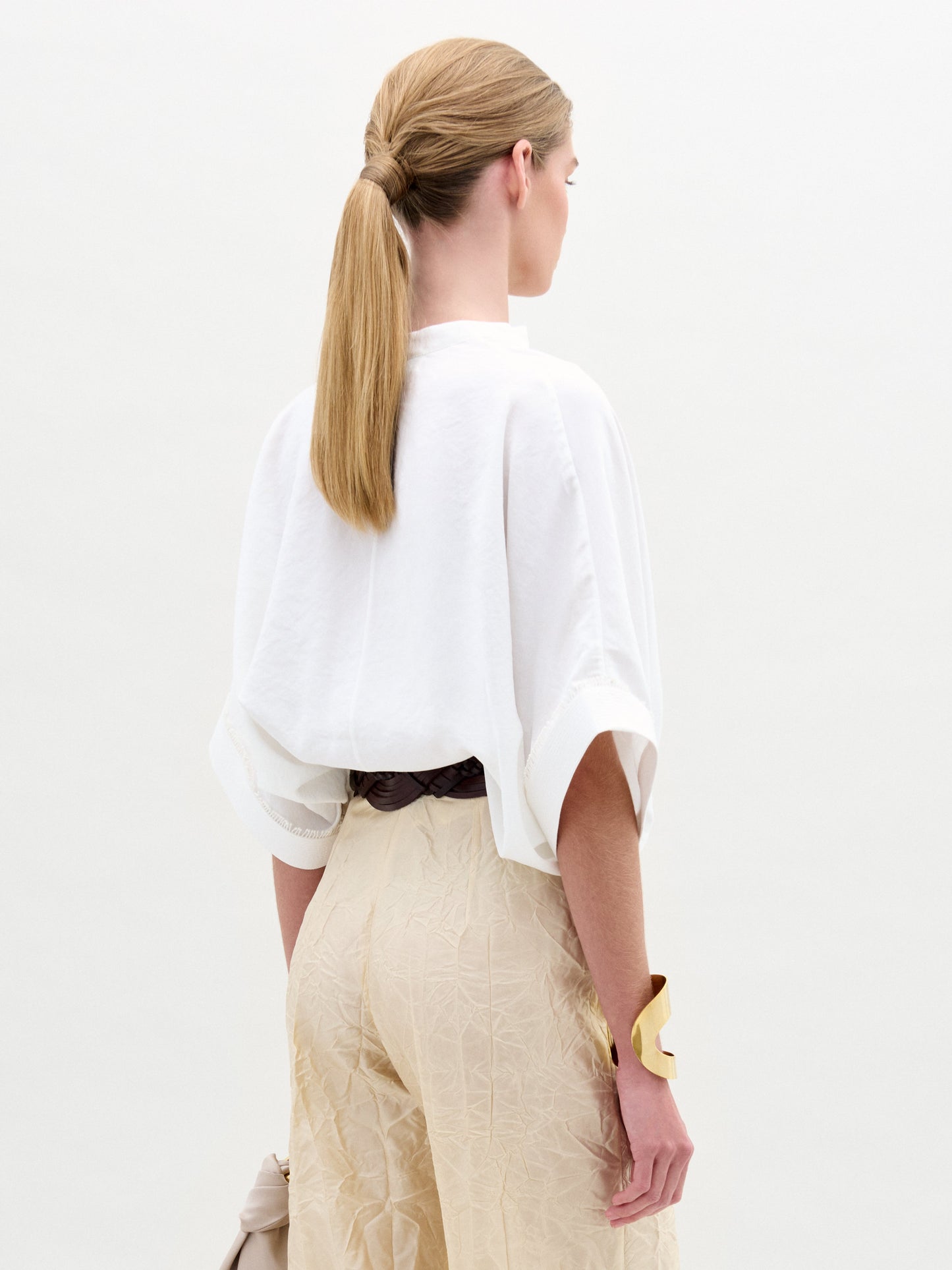 A person in a Daia Blouse paired with beige pants and a large belt stands against a plain background, accessorized at the wrist.