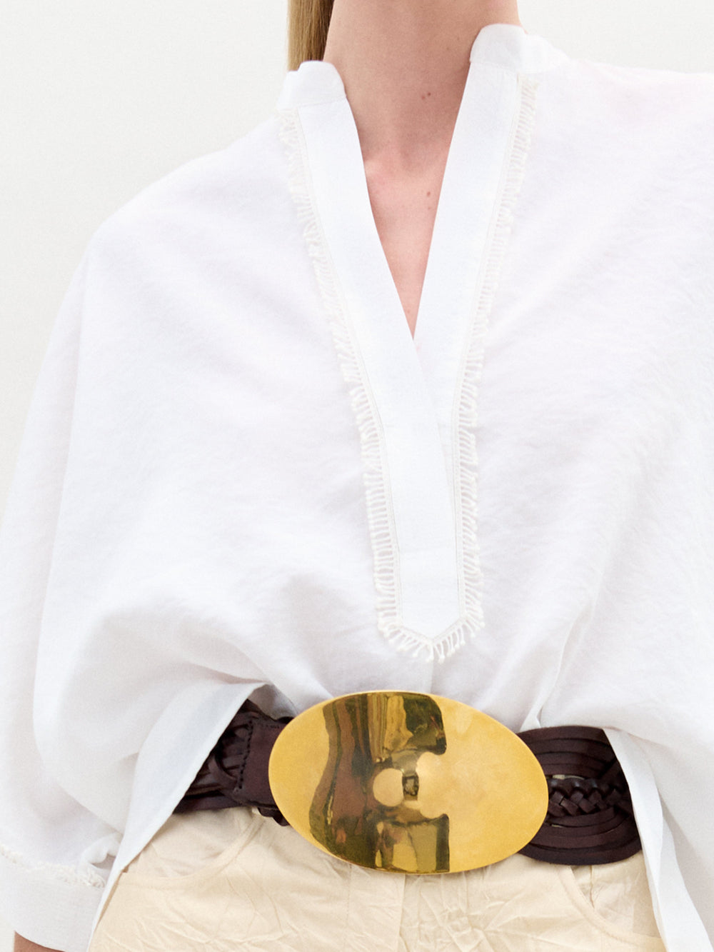 Una persona lleva puesta la Blusa Daia, una blusa blanca con textura y escote en V pronunciado, confeccionada con una exquisita tela italiana, complementada con un cinturón tejido de color marrón oscuro con una gran hebilla ovalada de oro. La imagen está recortada por encima del mentón de la persona.