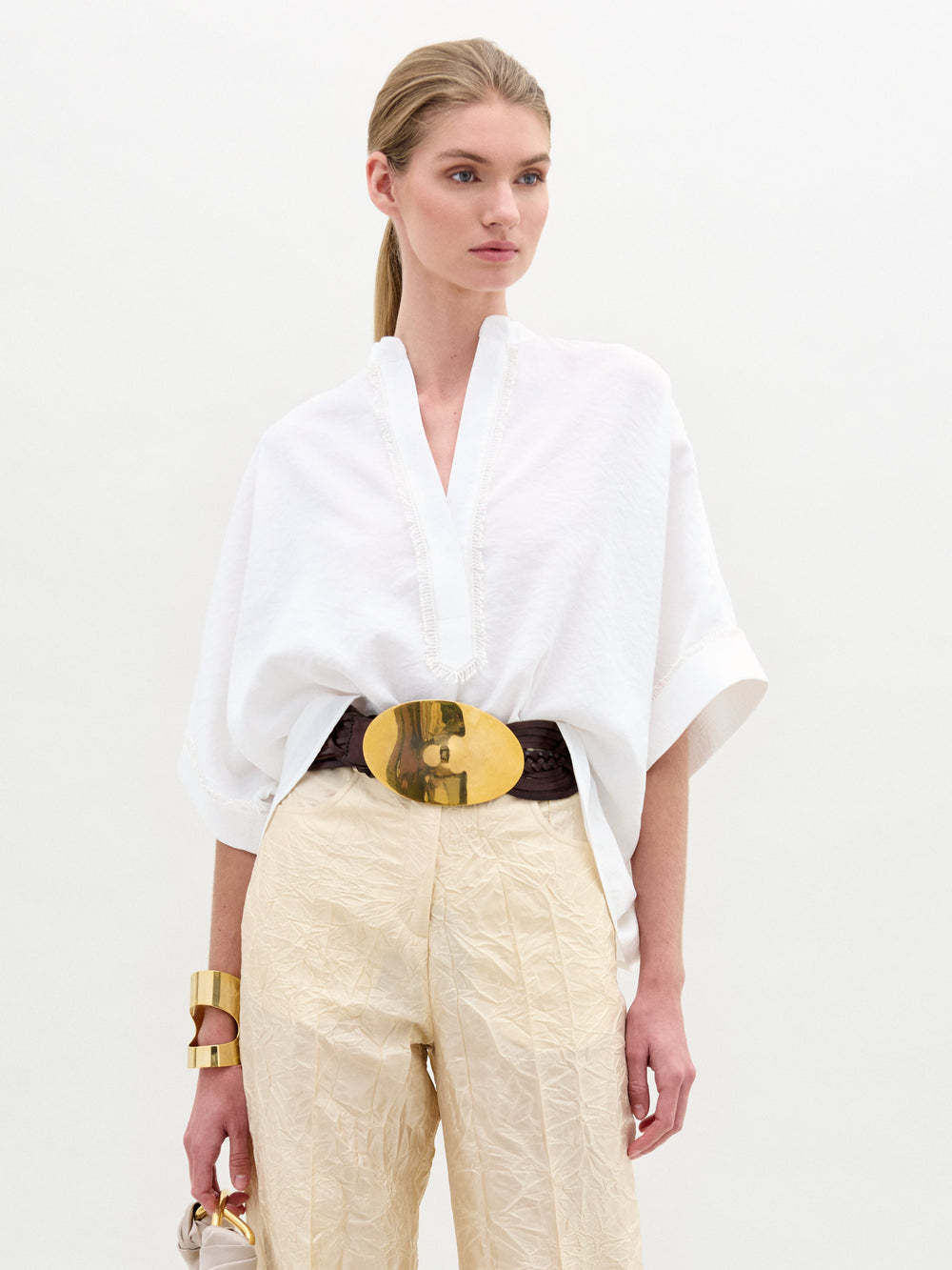 A person in a Daia Blouse paired with beige pants and a large belt stands against a plain background, accessorized at the wrist.