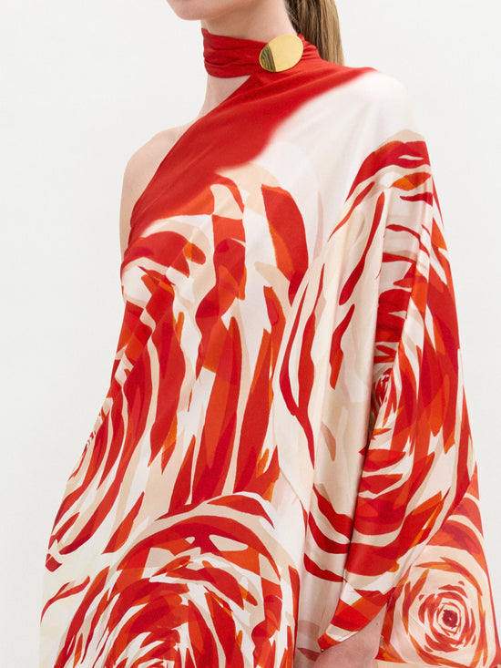 Con la Túnica Dani, un vestido asimétrico de un solo hombro en seda italiana con estampado floral rojo y blanco, con un drapeado elegante. Combinado con un accesorio de cuello rojo que presenta un gran detalle redondo dorado sobre un fondo blanco liso.