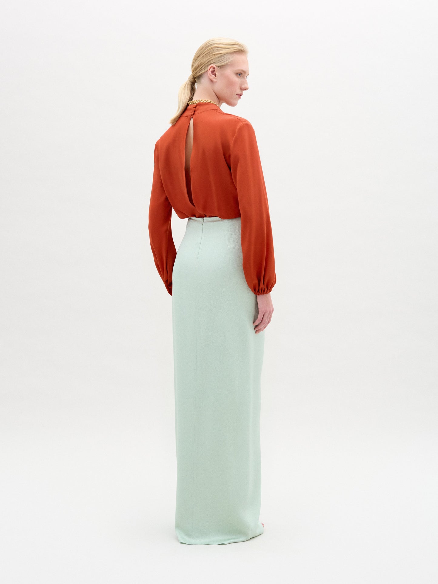 A person stands facing away, modeling the Delfina Blouse—a long-sleeved, burnt orange silk top with a keyhole back—paired with a high-waisted, floor-length, light mint green Italian fabric skirt against a white background.