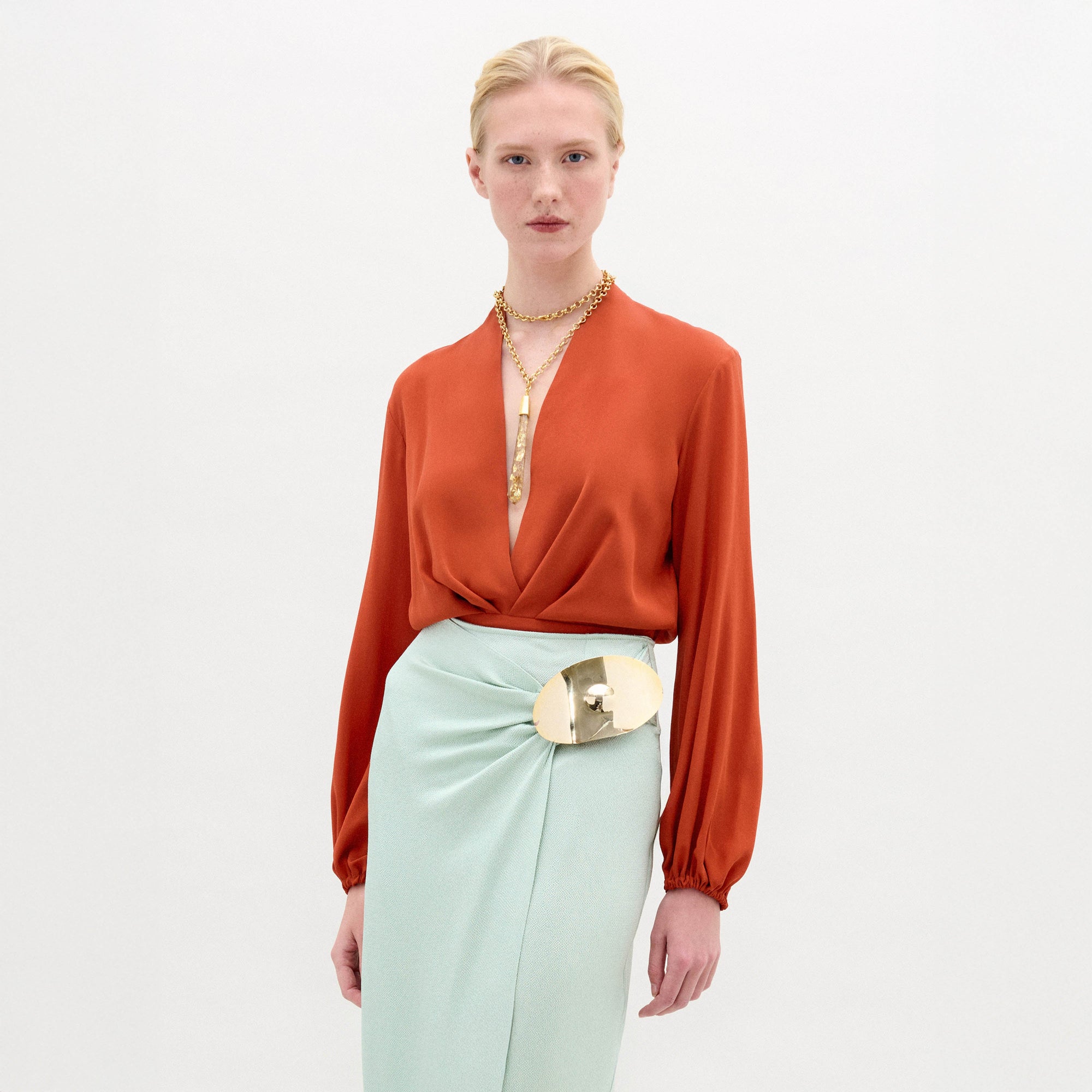 A woman stands against a plain light background wearing the Delfina Blouse, a long-sleeved deep orange silk top with a fitted waist, paired with a pale mint green skirt featuring a gold metallic belt and layered gold necklaces.