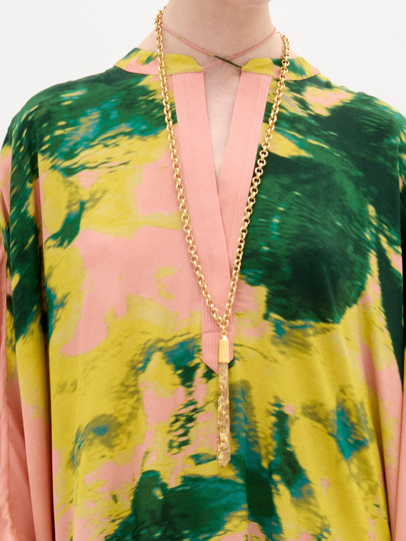 Vestidas con la Túnica Elea, una vibrante prenda 100% Seda con estampados en rosa, verde y amarillo, llevan un collar de cadena dorada con un dije texturizado. La parte superior tiene un escote en forma de V con una franja rosa sobre un sencillo fondo blanco.