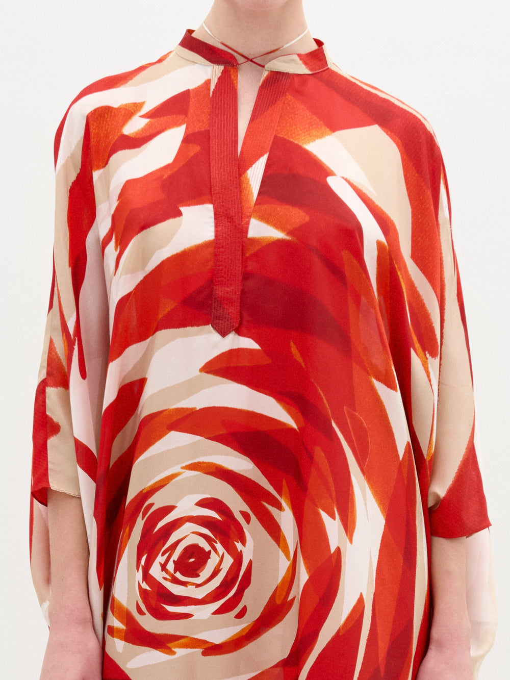 Alguien lleva puesta la Túnica Elea, una camisa colorida de manga larga hecha 100 % de seda, con un llamativo estampado abstracto en espiral en rojo y naranja. Tiene cuello mandarín, escote en forma de V y está colocada sobre un fondo claro y liso. No se ve el rostro de la persona.