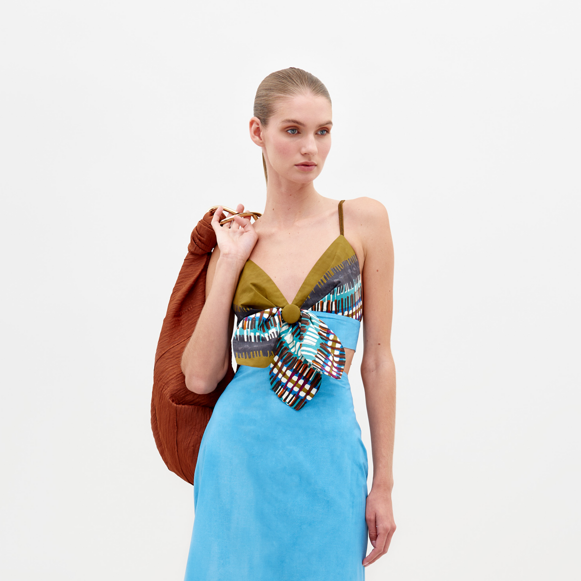 Una persona aparece de pie sobre un fondo liso con un vestido azul con un lazo estampado y un Top Flor, una blusa verde oliva con escote en V. Lleva un bolso grande con textura de color óxido sobre el hombro y el pelo recogido en un elegante moño.