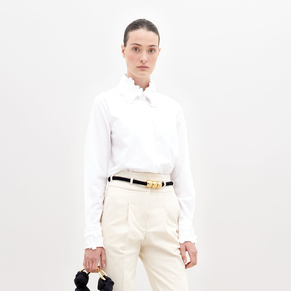 Una persona posa sobre un fondo blanco liso, vistiendo la Blusa Flower (una camisa blanca 100% algodón con apliques florales en el cuello y los puños), pantalones color crema y un cinturón negro, sosteniendo un objeto oscuro en una mano.
