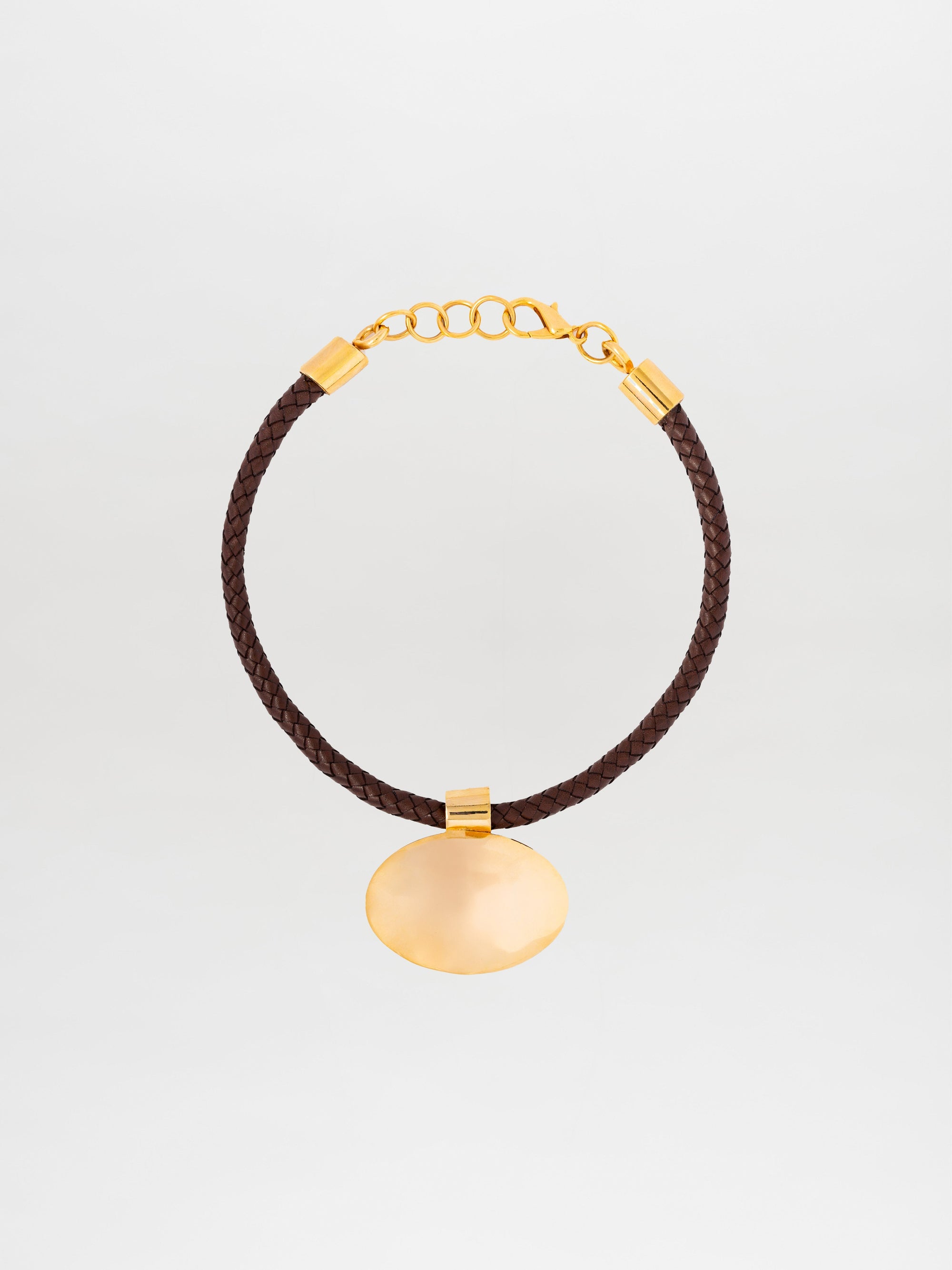 El Collar Gimena es una gargantilla de cuero trenzado marrón con cadena y broche de oro, elaborada en Colombia. Presenta un gran colgante ovalado de oro en el centro, sobre un fondo liso de color claro.