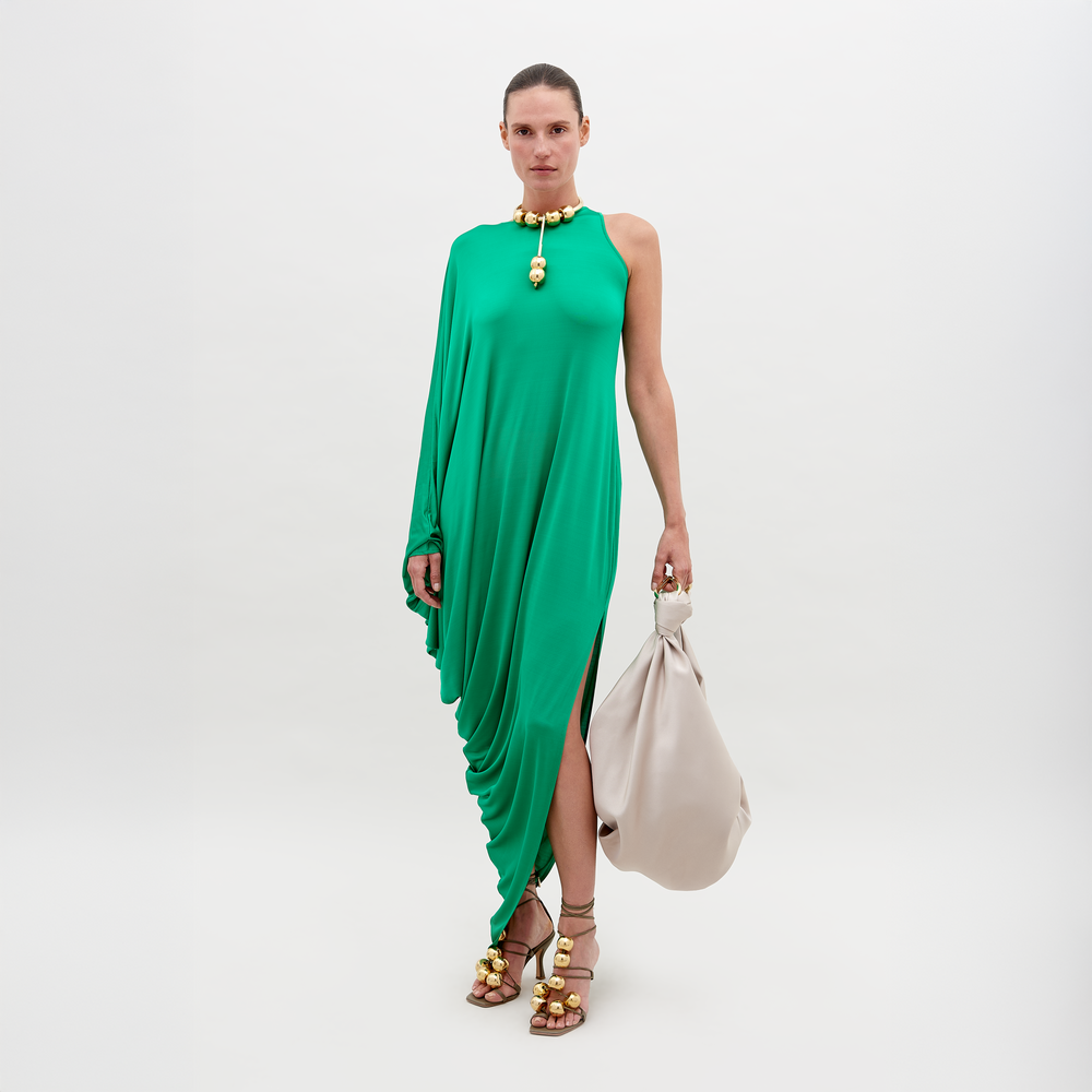 Una persona está de pie con un Vestido Golda verde de una sola manga con efecto drapeado, sosteniendo un gran bolso beige y usando sandalias marrones con punta abierta y adornos dorados sobre un fondo blanco liso.
