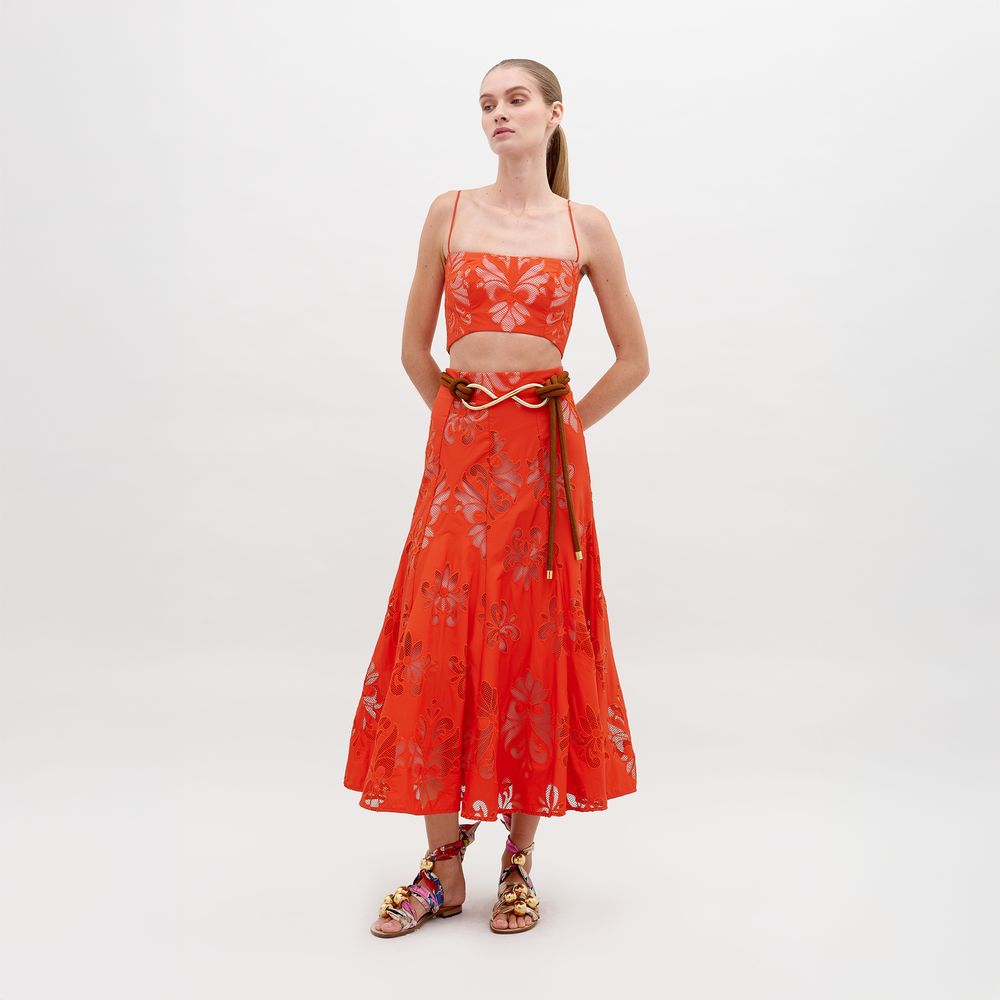 Una persona está parada sobre un fondo blanco liso con un top corto con estampado floral de colores brillantes de Falda Grettel y una falda midi a juego. Junta sus manos al frente y completa su look con unas sandalias de tiras coloridas confeccionadas con tela italiana de primera calidad.