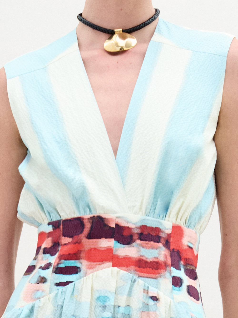 Una persona lleva el Vestido Jocelyn, un vestido midi sin mangas con escote en V profundo, fabricado en Colombia. Tiene una parte superior de rayas celestes y blancas y una falda con estampado abstracto en rojo, azul y blanco. Completa el look con una gargantilla negra con un colgante dorado.