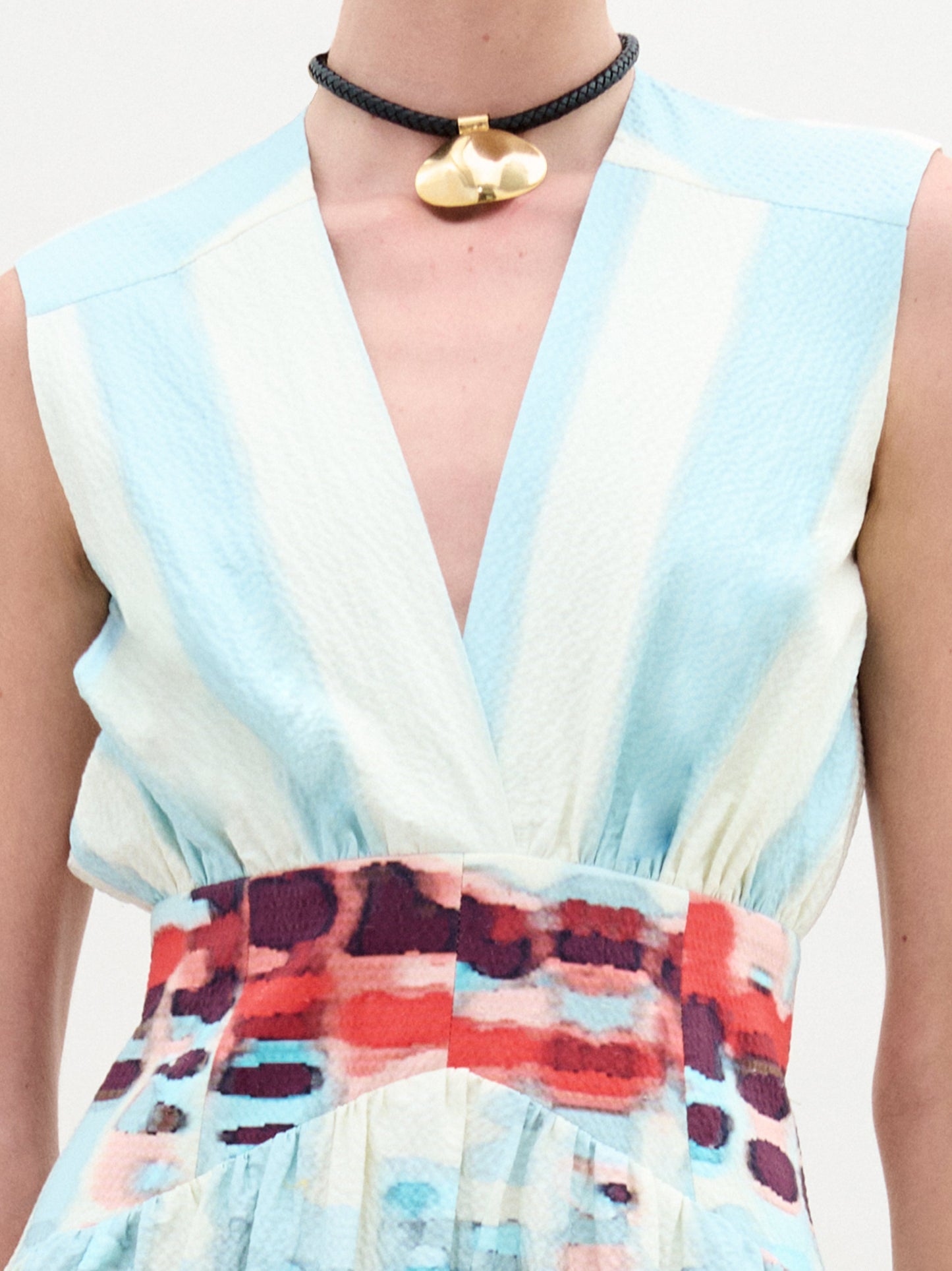 Una persona lleva el Vestido Jocelyn, un vestido midi sin mangas con escote en V profundo, fabricado en Colombia. Tiene una parte superior de rayas celestes y blancas y una falda con estampado abstracto en rojo, azul y blanco. Completa el look con una gargantilla negra con un colgante dorado.