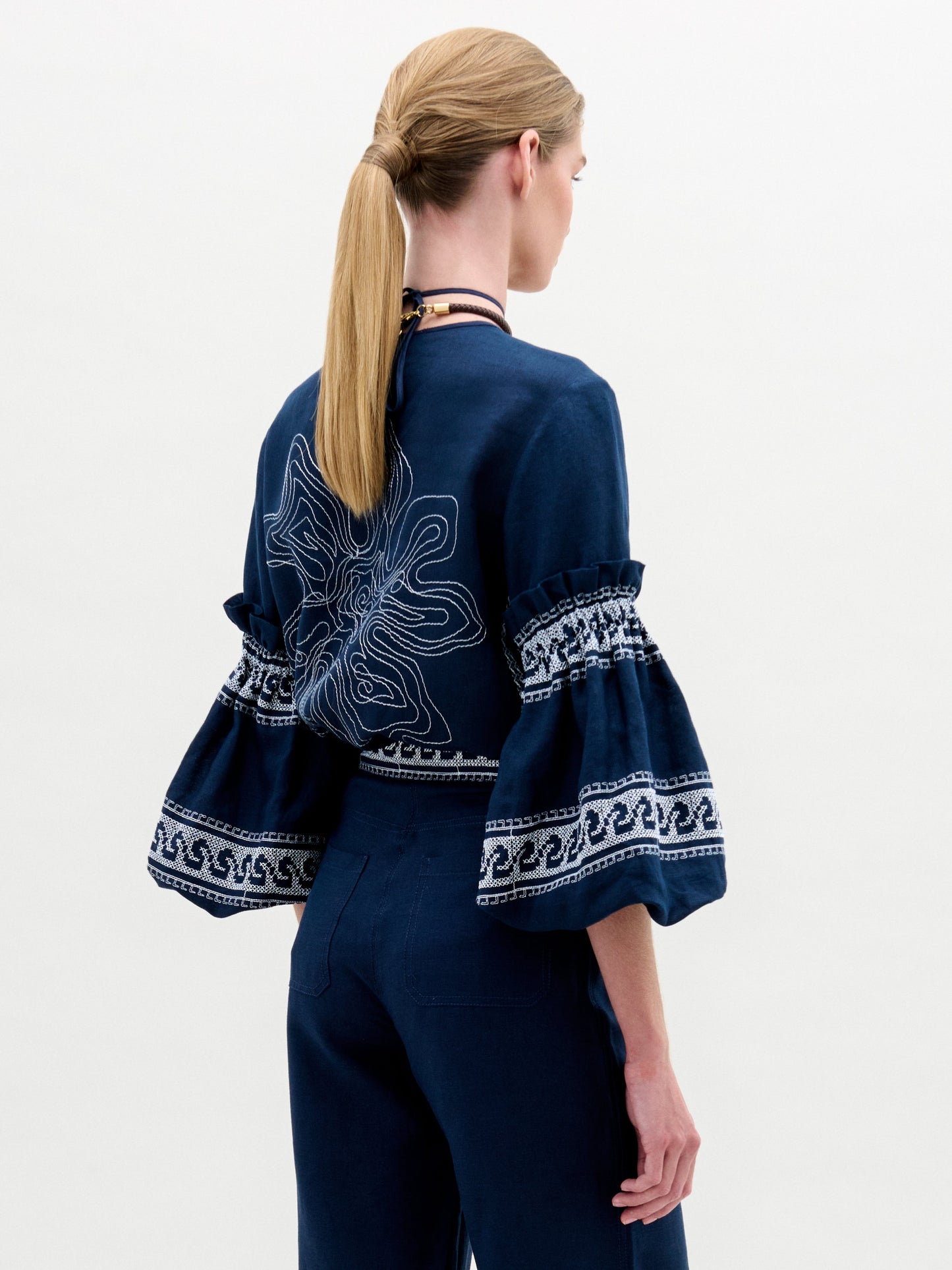 A woman models the Lucaya Blouse, showcasing its intricate white patterns and ruffled sleeves, paired elegantly with a turquoise necklace against a simple background.