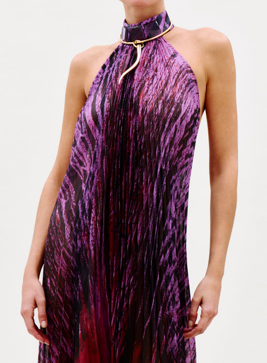 A woman poses against a plain light background wearing the Lumi Dress, a sleeveless purple and black patterned piece paired with a high collar necklace and holding a bright pink clutch.