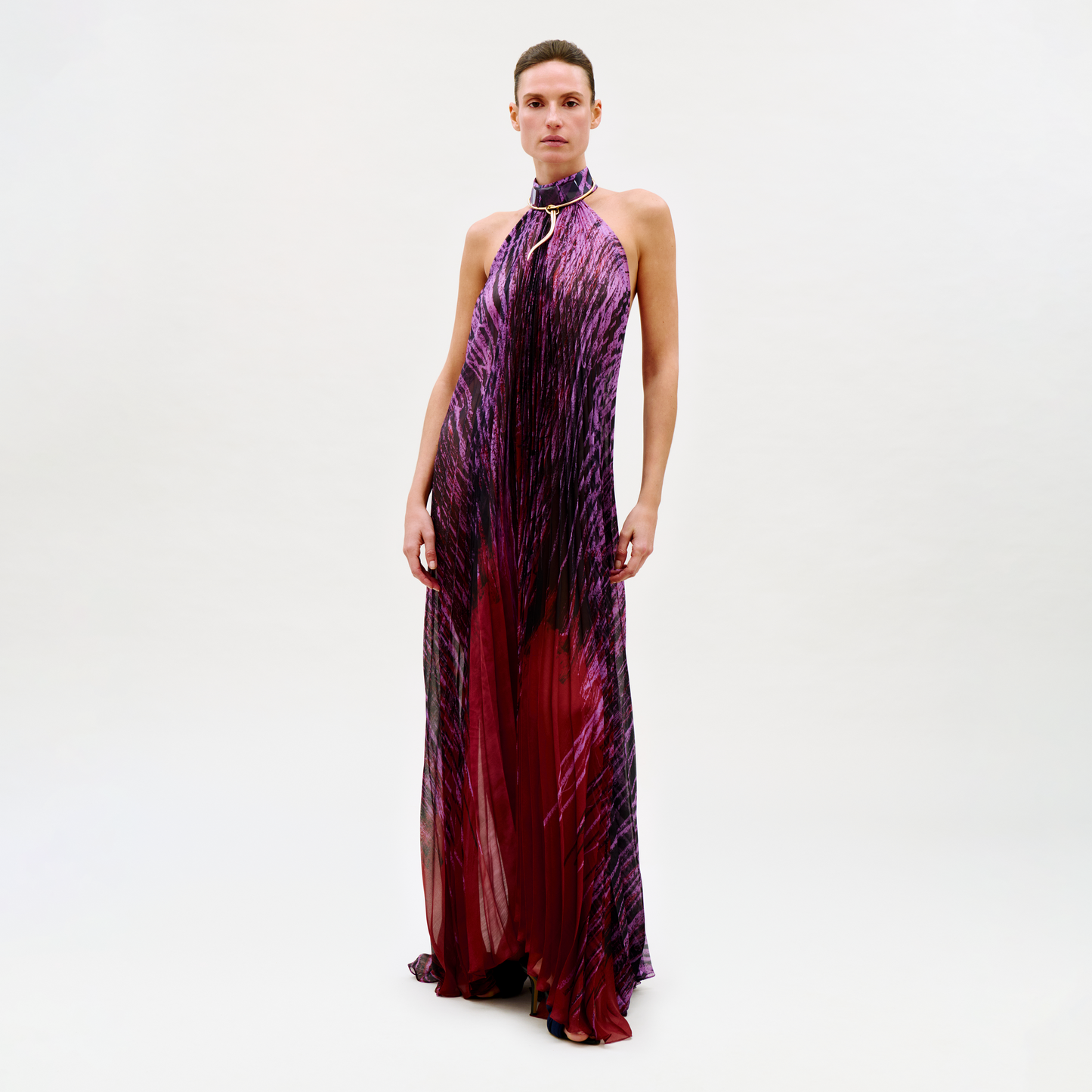 A woman poses against a plain background in the sleeveless, floor-length Lumi Dress, which features a purple and red gradient, halter neck, keyhole neckline, and gold accent detail.