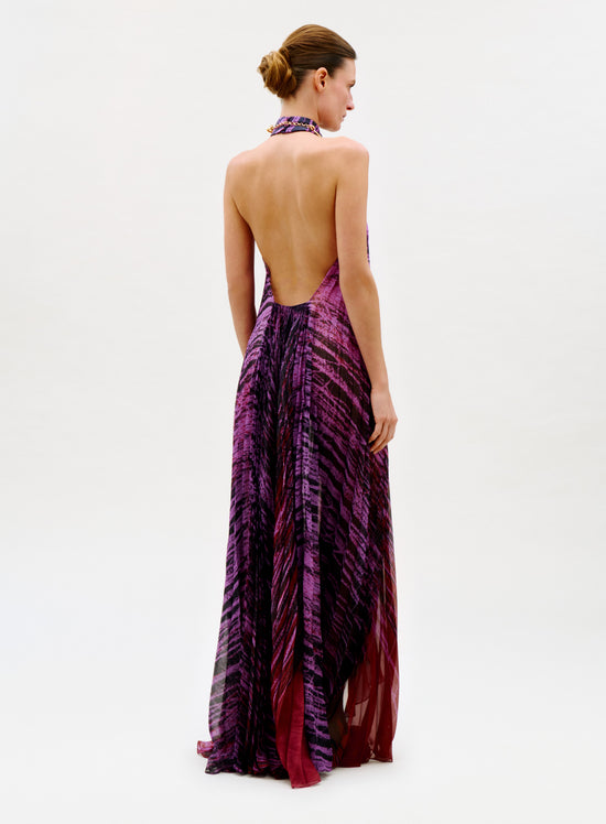 Wearing the Lumi Dress—a long, flowing, backless halter dress in purple and magenta abstract patterns—a woman stands with her back to the camera. Her hair is in a neat bun against a plain white background.