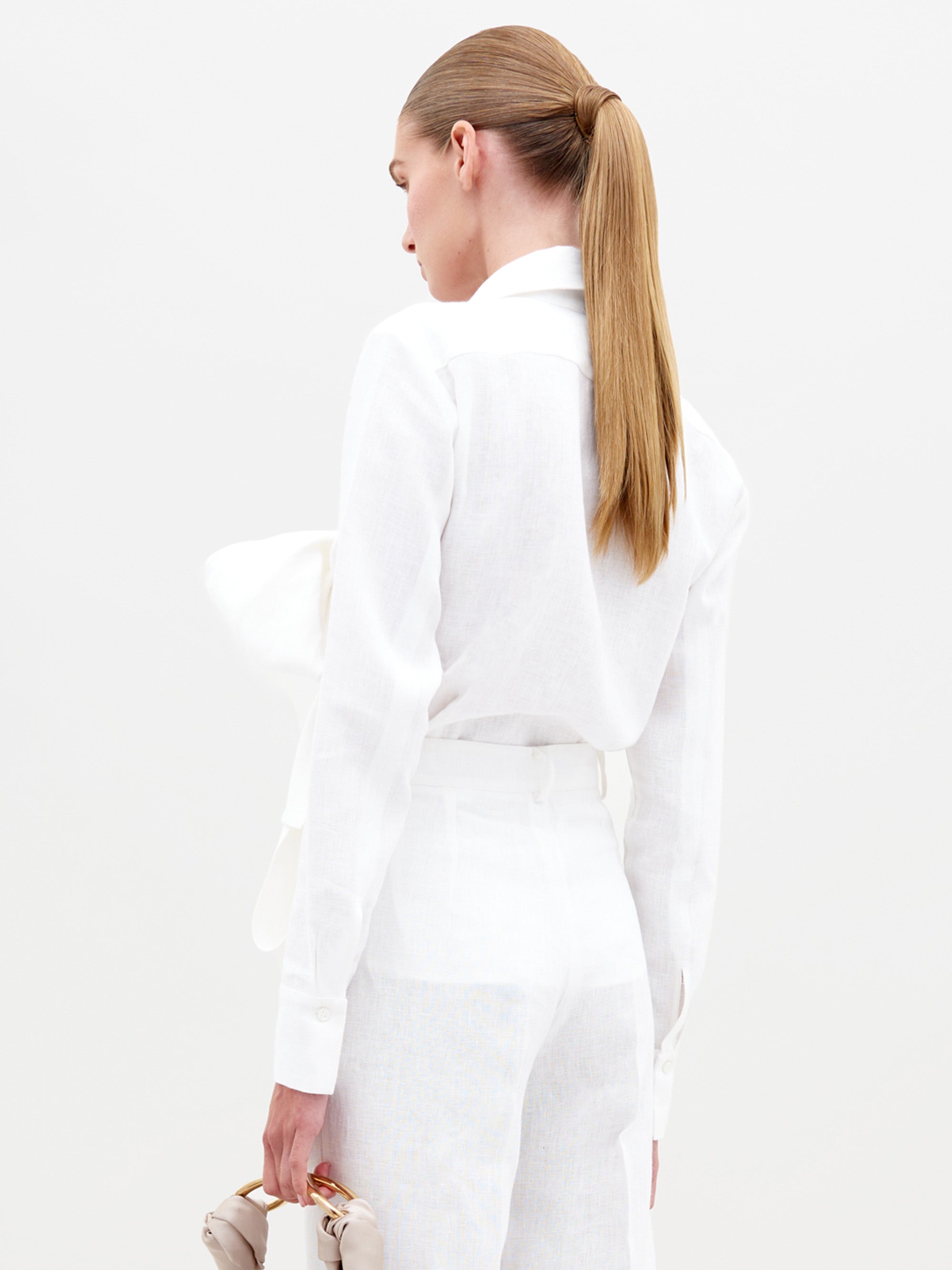A person with long light brown hair in a ponytail is wearing the Mercury Blouse (Linen) White with a large bow, white pants, and holding beige shoes. Pre-order now to ensure you get this look by the 2025 ship-by date.