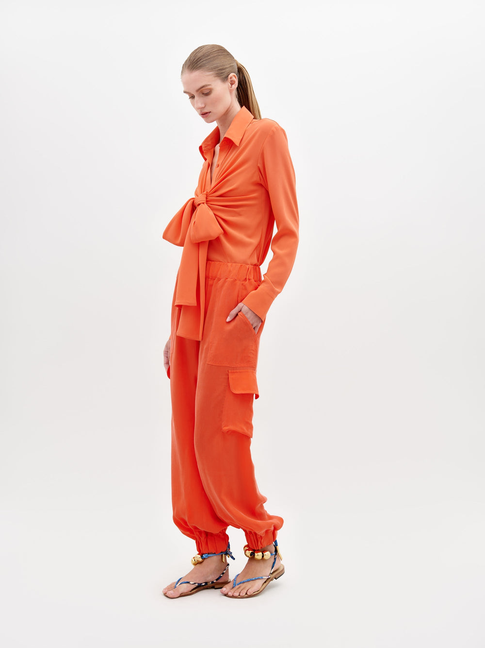 Sobre un fondo blanco, una persona lleva un conjunto naranja confeccionado con un exquisito textil italiano. El conjunto incluye una camiseta de manga larga con un gran lazo y pantalones cargo Pantalón Jess a juego con bolsillos. Unas sandalias decorativas completan el elegante look.