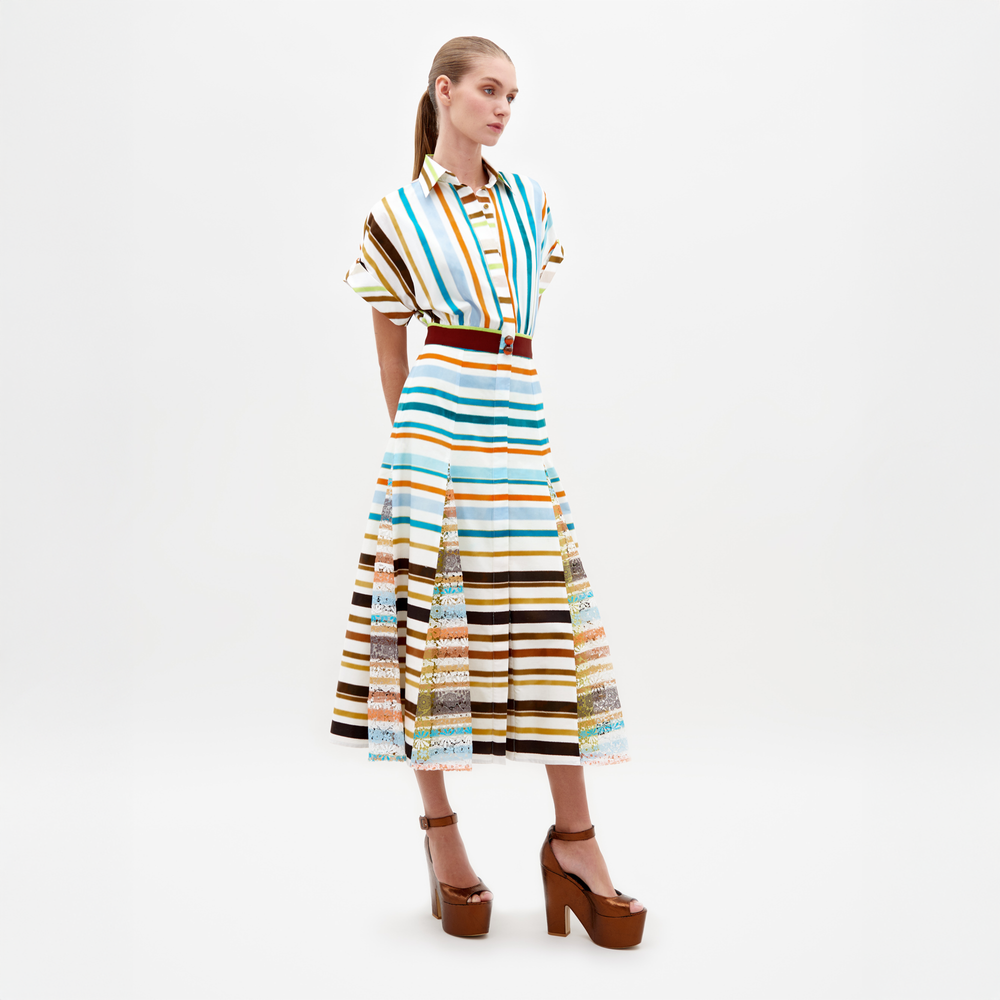 Una persona está de pie con el Vestido Odila, un vestido midi con estampado de rayas multicolores en azul, naranja, amarillo, marrón y blanco. Tiene una cintura ajustada con cinturón y llega por debajo de las rodillas. Lo combina con tacones de plataforma marrones y tiene el pelo largo y liso sobre un fondo blanco liso.