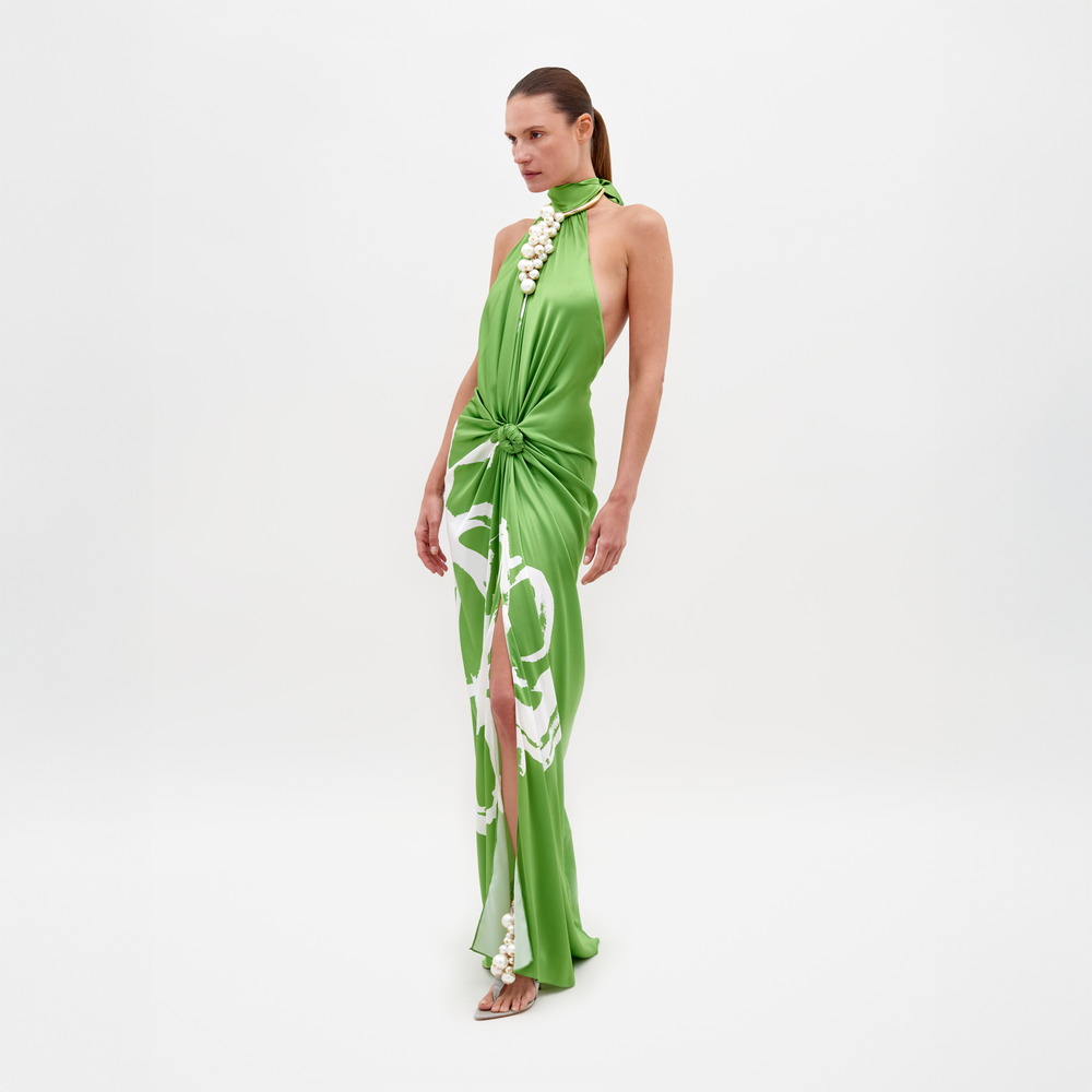 Sobre un fondo blanco, una mujer lleva el Vestido Olatz, un vestido largo sin mangas de seda verde brillante con un estampado floral abstracto. Presenta un diseño de cuello halter, abertura alta y detalle de cintura fruncida. El look se completa con sandalias de tacón alto y cabello largo recogido.
