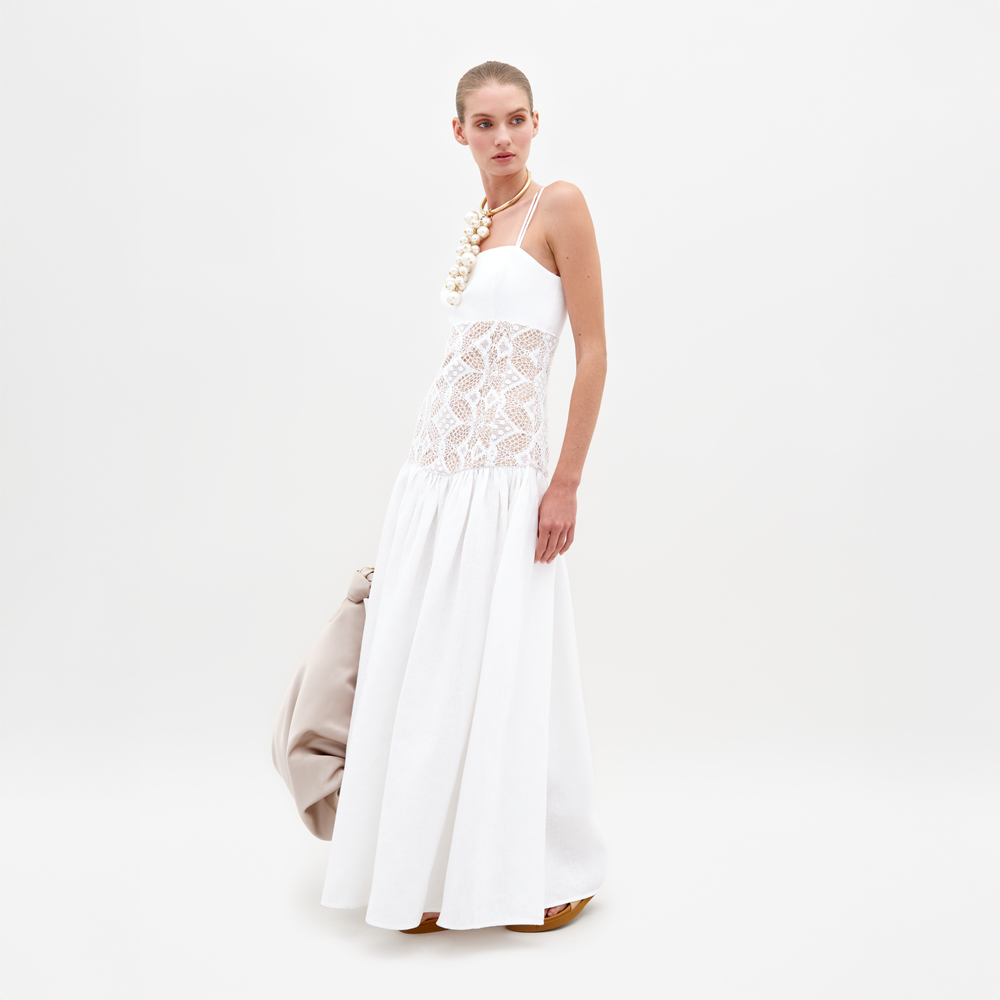 Una persona con un Vestido Pilar blanco con intrincados detalles de encaje sostiene un bolso beige y lleva un collar distintivo con cuentas grandes, todo ello sobre un fondo blanco liso.