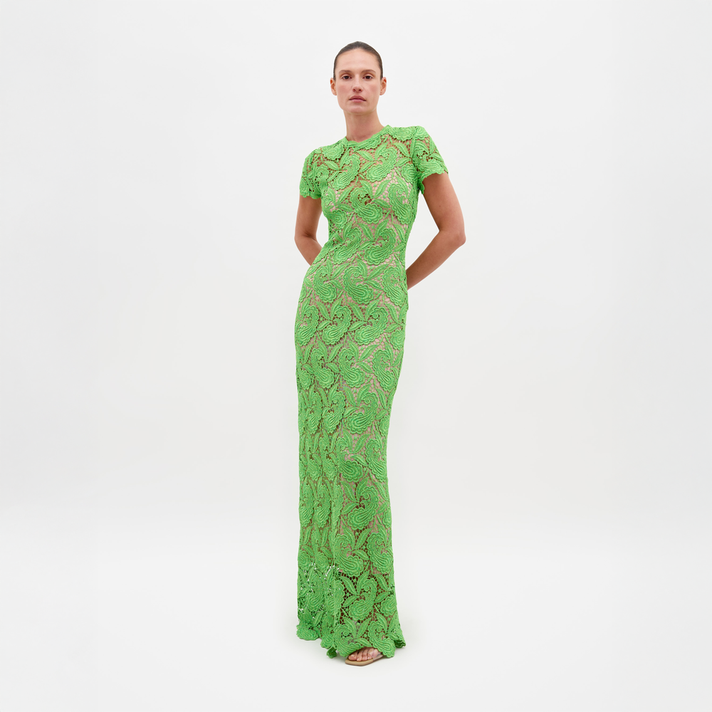Una persona con un vestido Roxana de encaje verde largo y ajustado con mangas cortas está parada sobre un fondo blanco liso, mirando hacia adelante con las manos colocadas detrás de la espalda.