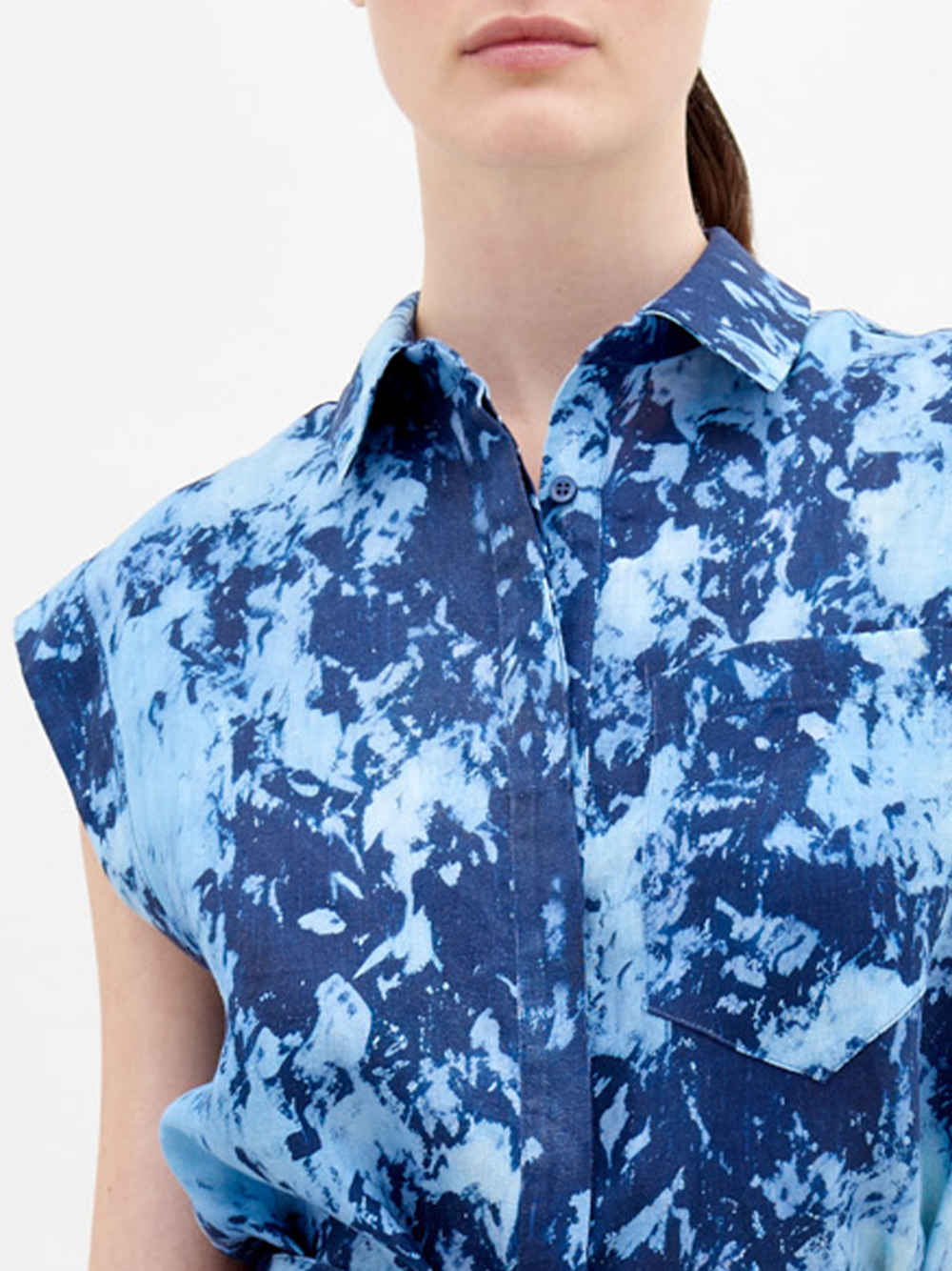 A woman wearing a blue tie-dye dress with a collar and sleeveless design, carrying a large navy blue bag on her shoulder, with her hair tied back in a ponytail, is showcasing the Rina Blouse Indigo exclusive pre-order item available until November 15th, 2024.
