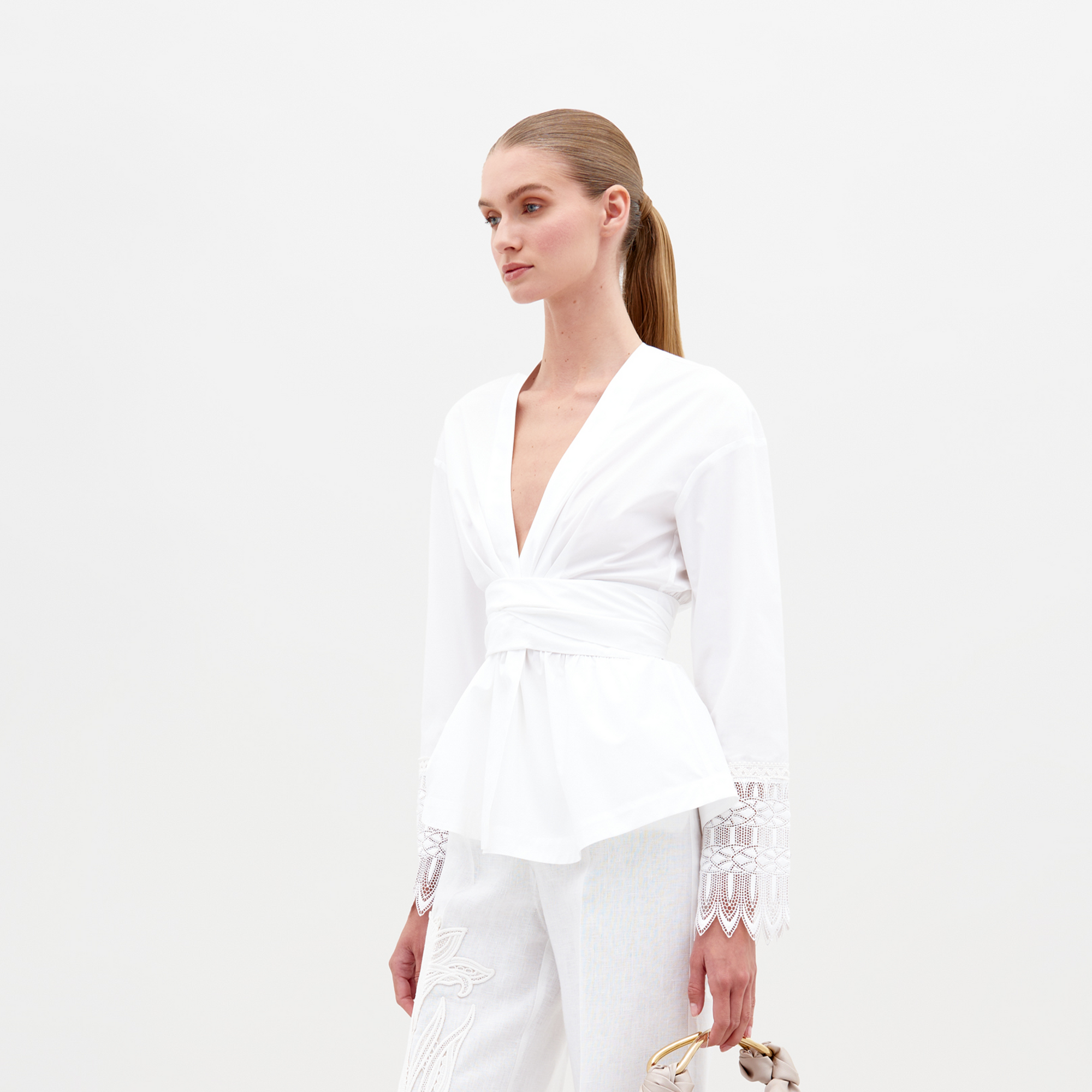 Una persona con el pelo largo atado hacia atrás lleva una blusa Saanvi blanca con escote en V y mangas de encaje y pantalones a juego. Sostiene una pequeña bolsa de color claro sobre un fondo blanco liso.