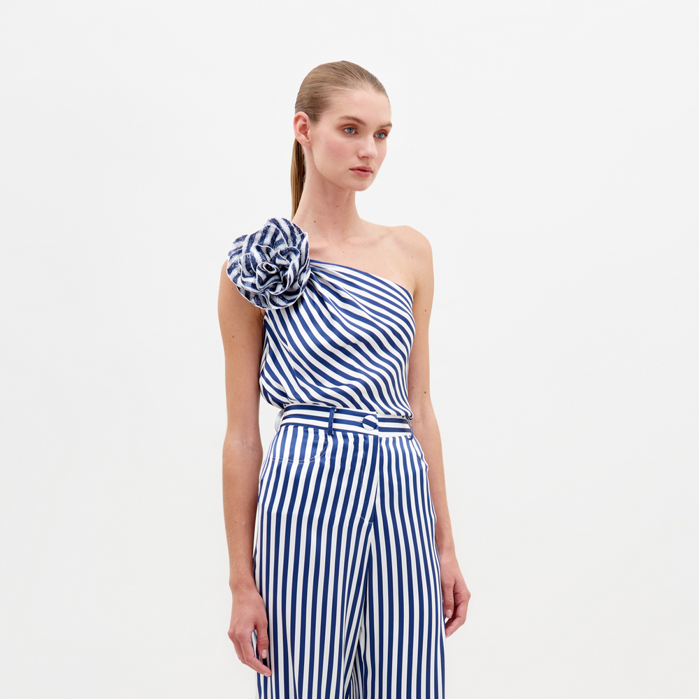 Una persona con cabello largo viste la Blusa Sebastiana, una blusa asimétrica de rayas azules y blancas con un solo hombro y un gran motivo floral en el hombro, combinada con pantalones de cintura alta a juego sobre un fondo blanco liso.