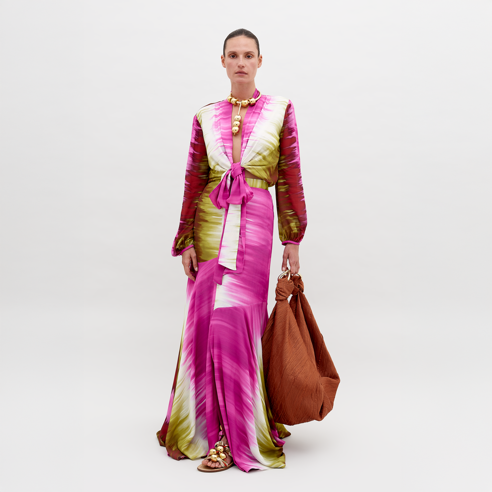 Una persona está de pie con la Falda Shane, un vestido largo con efecto tie-dye en tonos rosa, verde y blanco, con mangas largas y un lazo en la cintura. Sostiene una gran bolsa marrón y usa sandalias doradas y un collar dorado grueso sobre un fondo blanco liso.