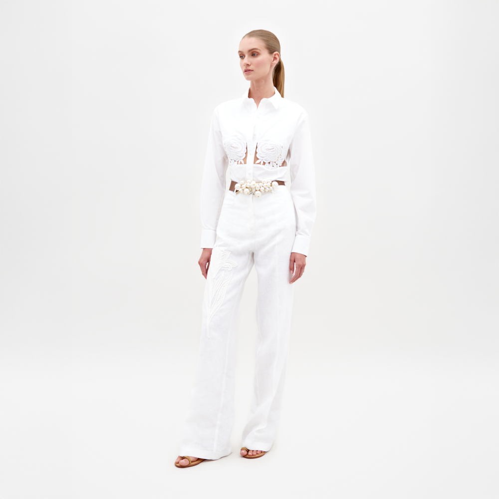 Sobre un fondo blanco liso, una persona lleva un traje blanco con una blusa con cortes elaborados y mangas largas con apliques florales. Los pantalones Pantalón Trilce, holgados y de cintura alta, se combinan con sandalias abiertas y un cinturón detallado.
